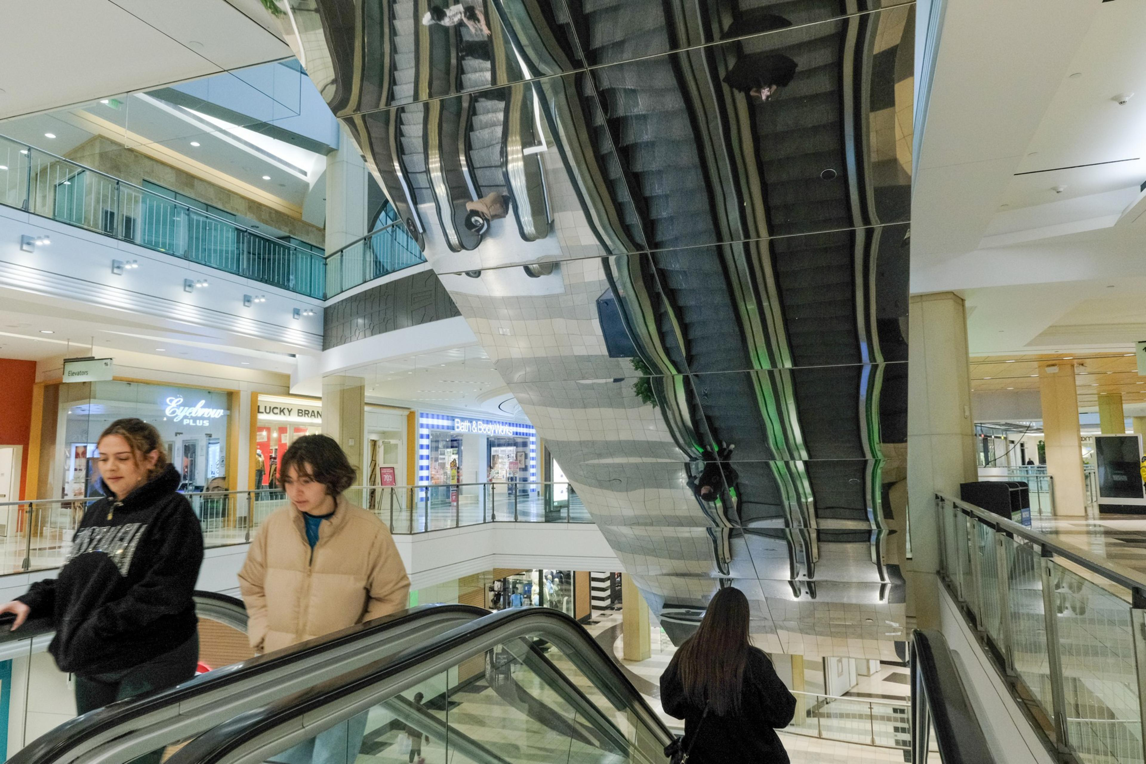 People riding an escalator in a mall with reflective ceiling creating mirrored images.