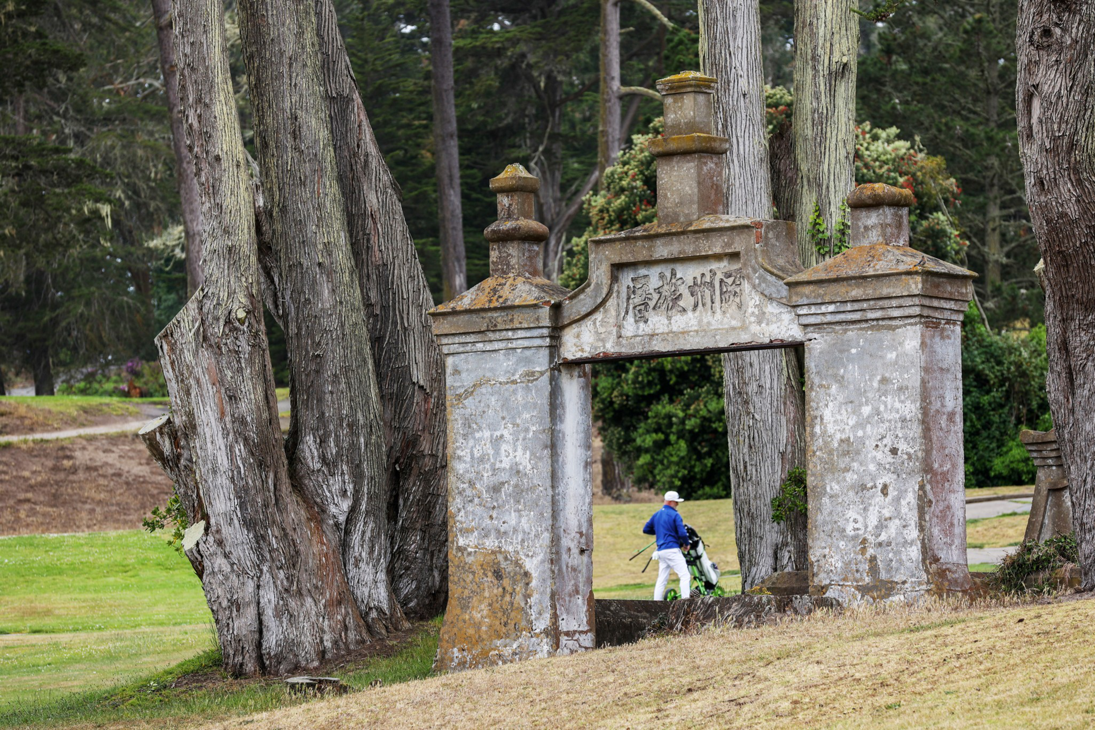 An old stone archway with weathered inscriptions stands among large trees on grassy ground, with a person pushing a golf bag in the background.