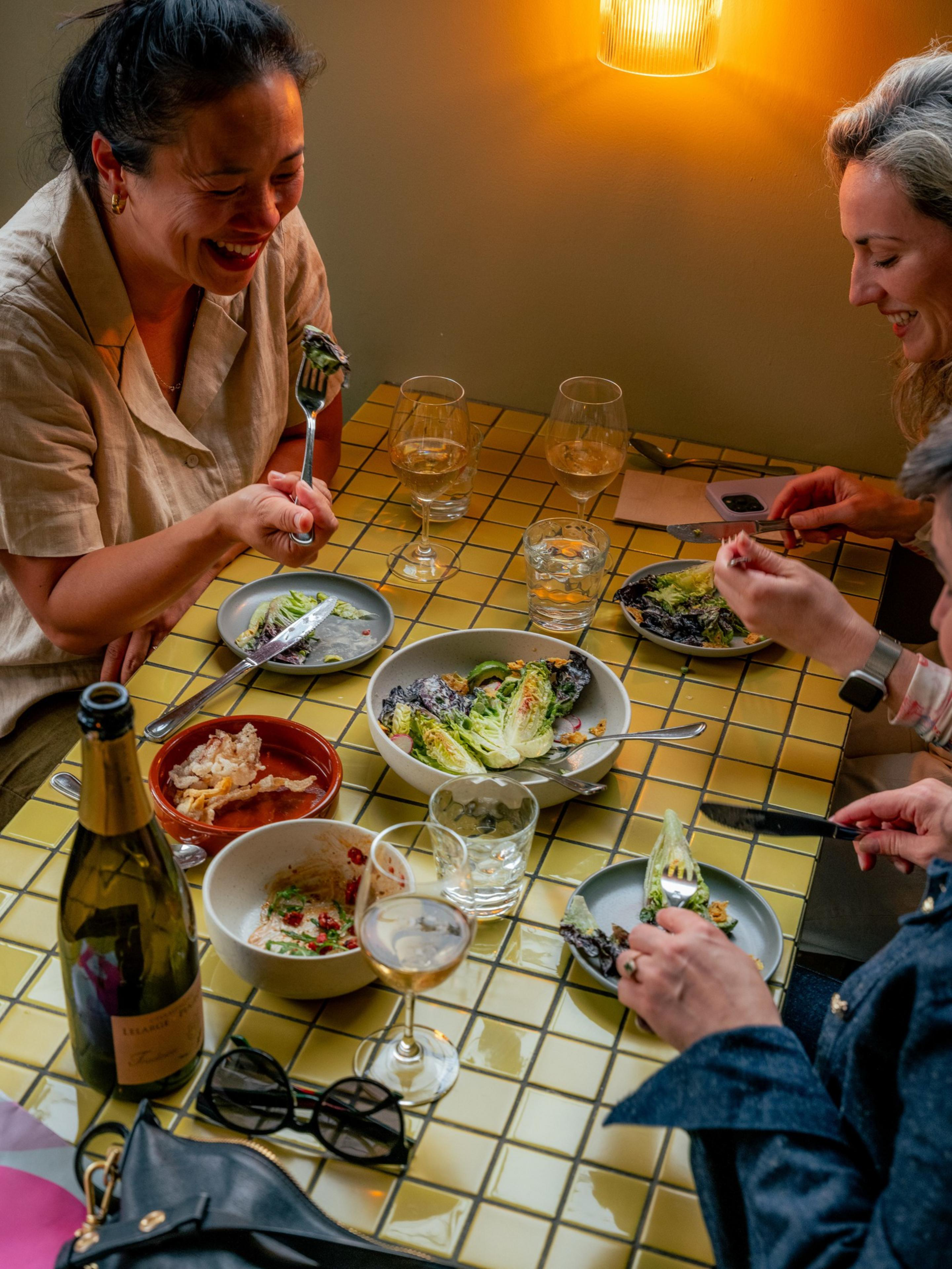 Four people are enjoying a meal at a table with plates of salad, drinks, and bowls of food. They are smiling and chatting under warm lighting.