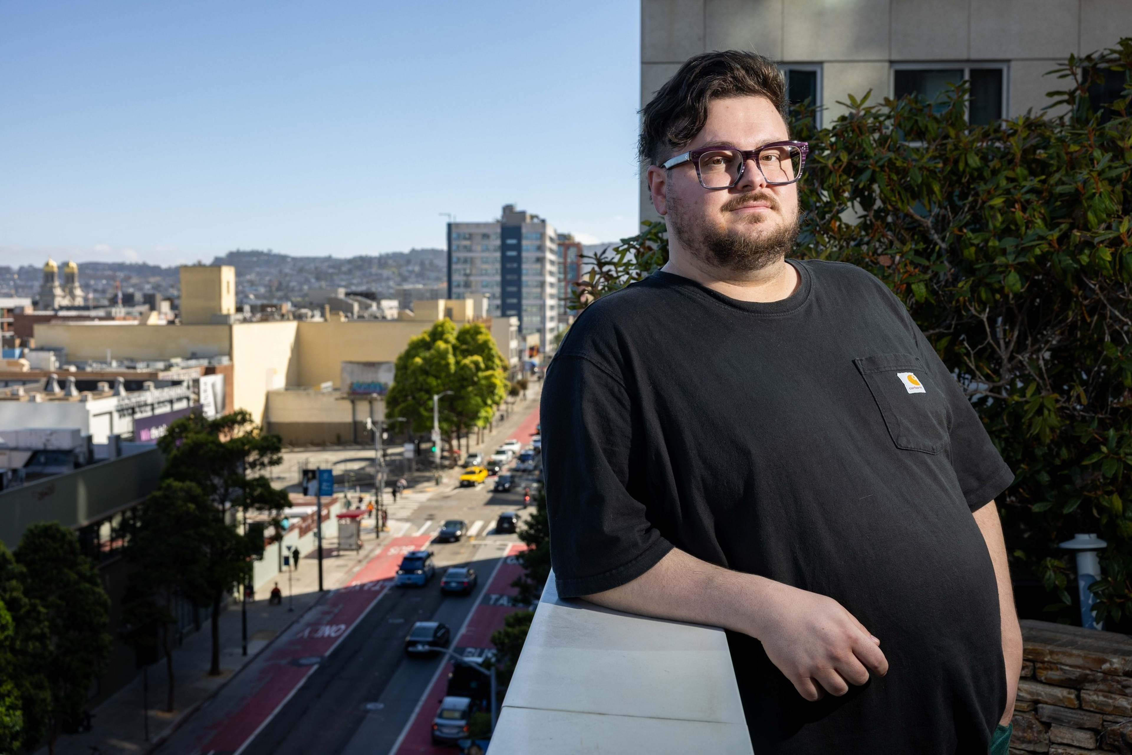 A man wearing glasses and a black T-shirt stands on a balcony overlooking a sunny urban street with cars and tall buildings.