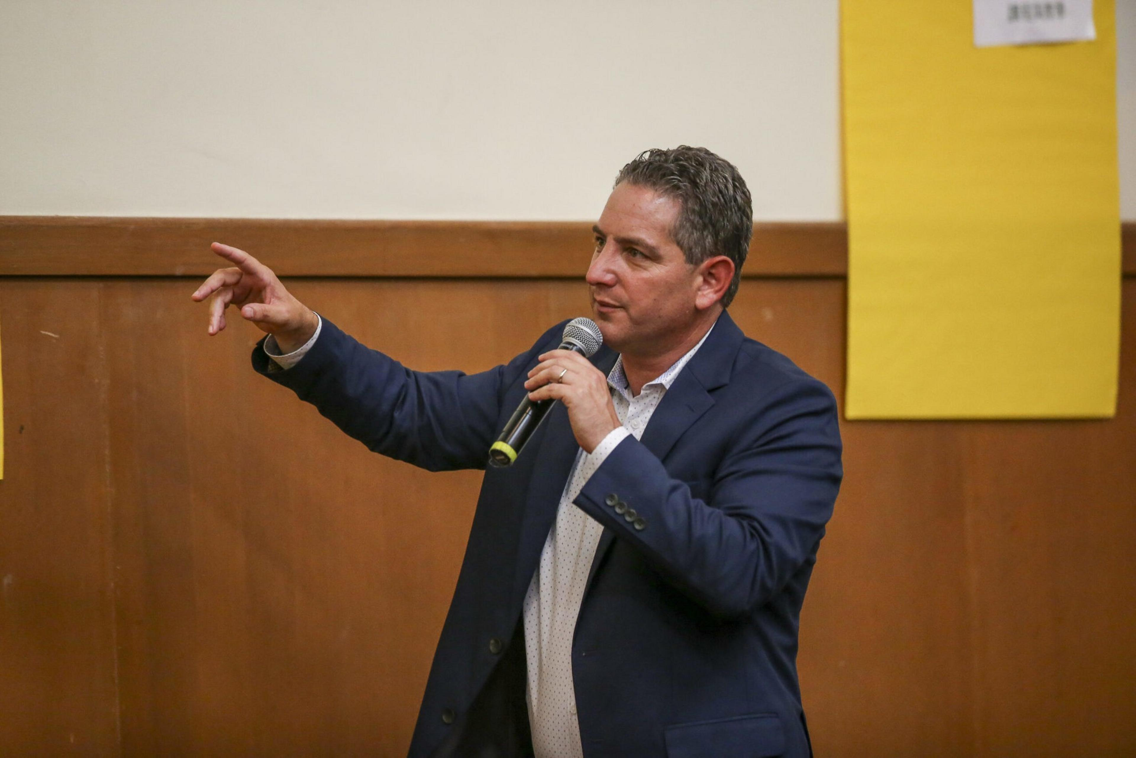 A man in a navy blazer and white shirt holds a microphone in one hand and gestures with the other while speaking indoors.