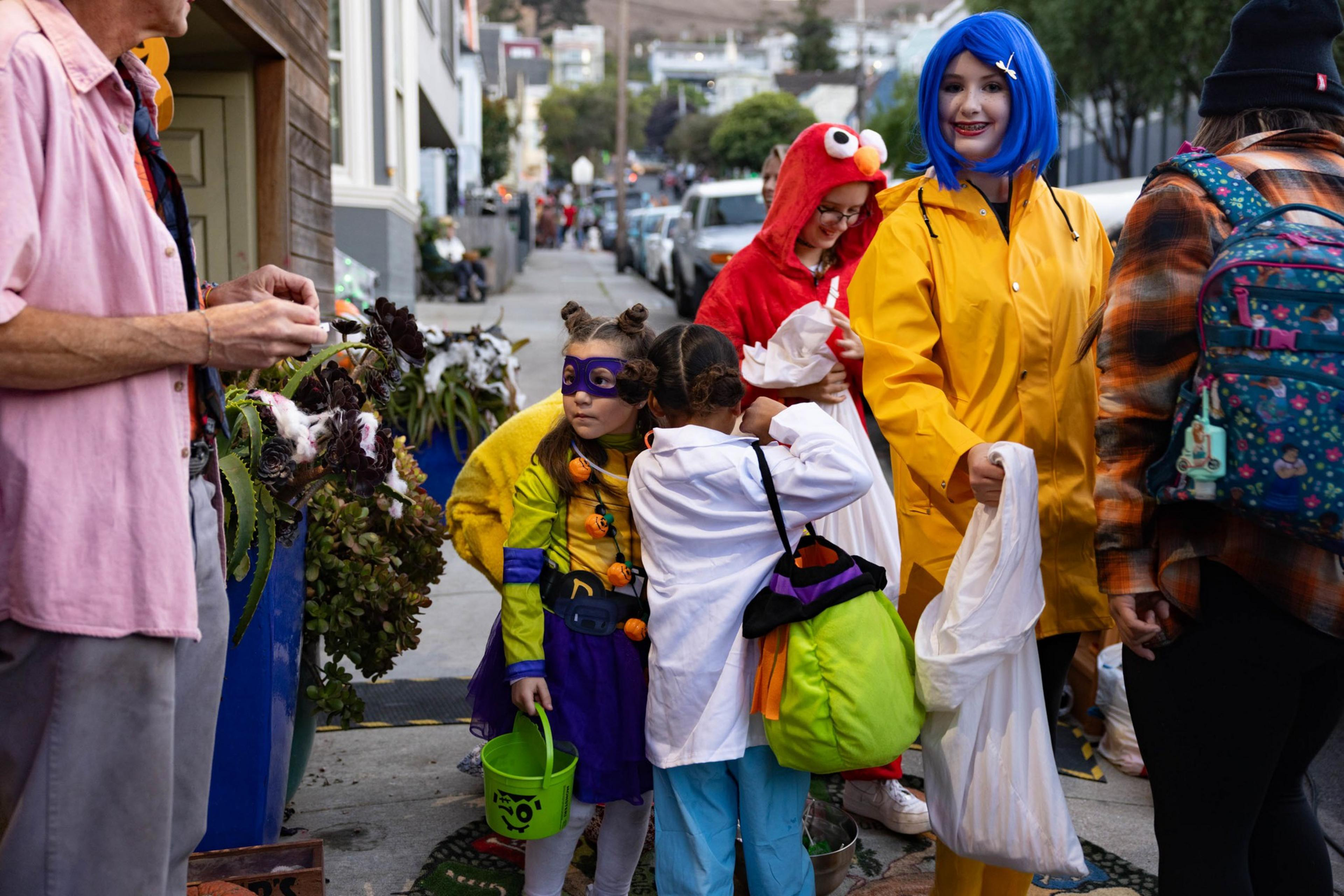 Children wearing Halloween costumes, including a superhero, a red monster, and a character in a yellow raincoat with blue hair, gather for trick-or-treating.