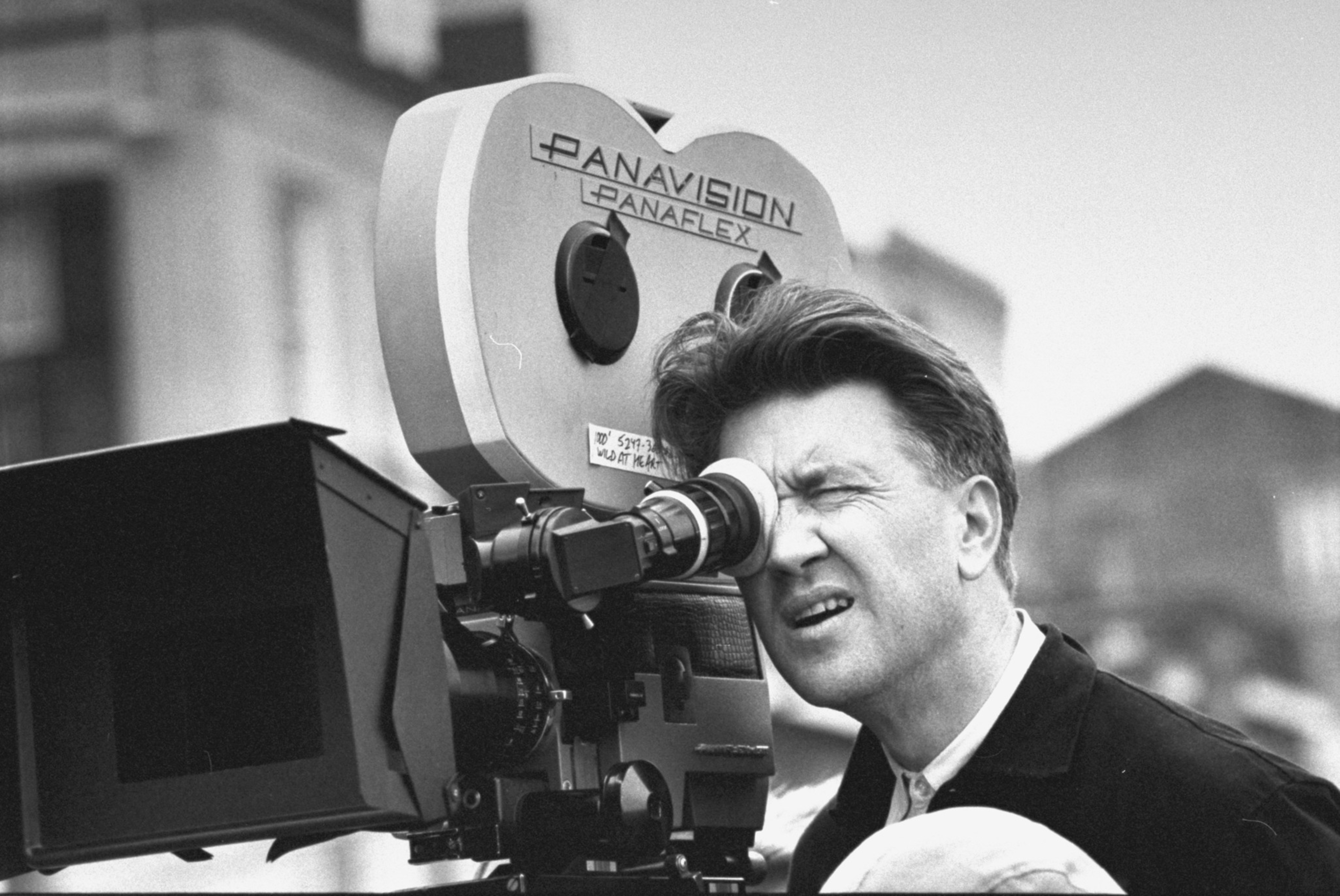 A person is looking through the viewfinder of a large Panavision movie camera outdoors, with a blurred building in the background. The scene is in black and white.