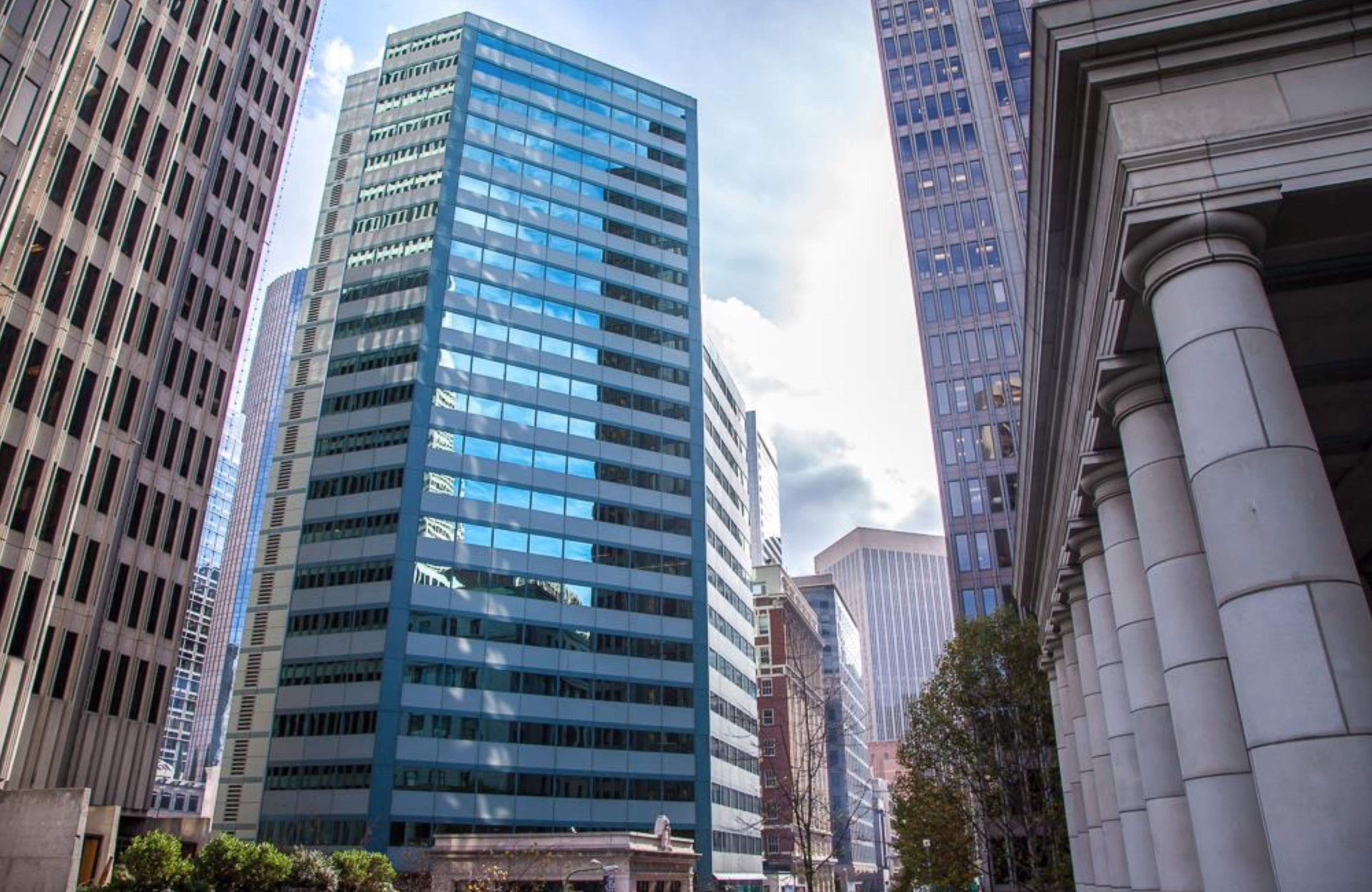 A cluster of modern city skyscrapers towers over the street. The central building has reflective glass, while columns and trees line the foreground.