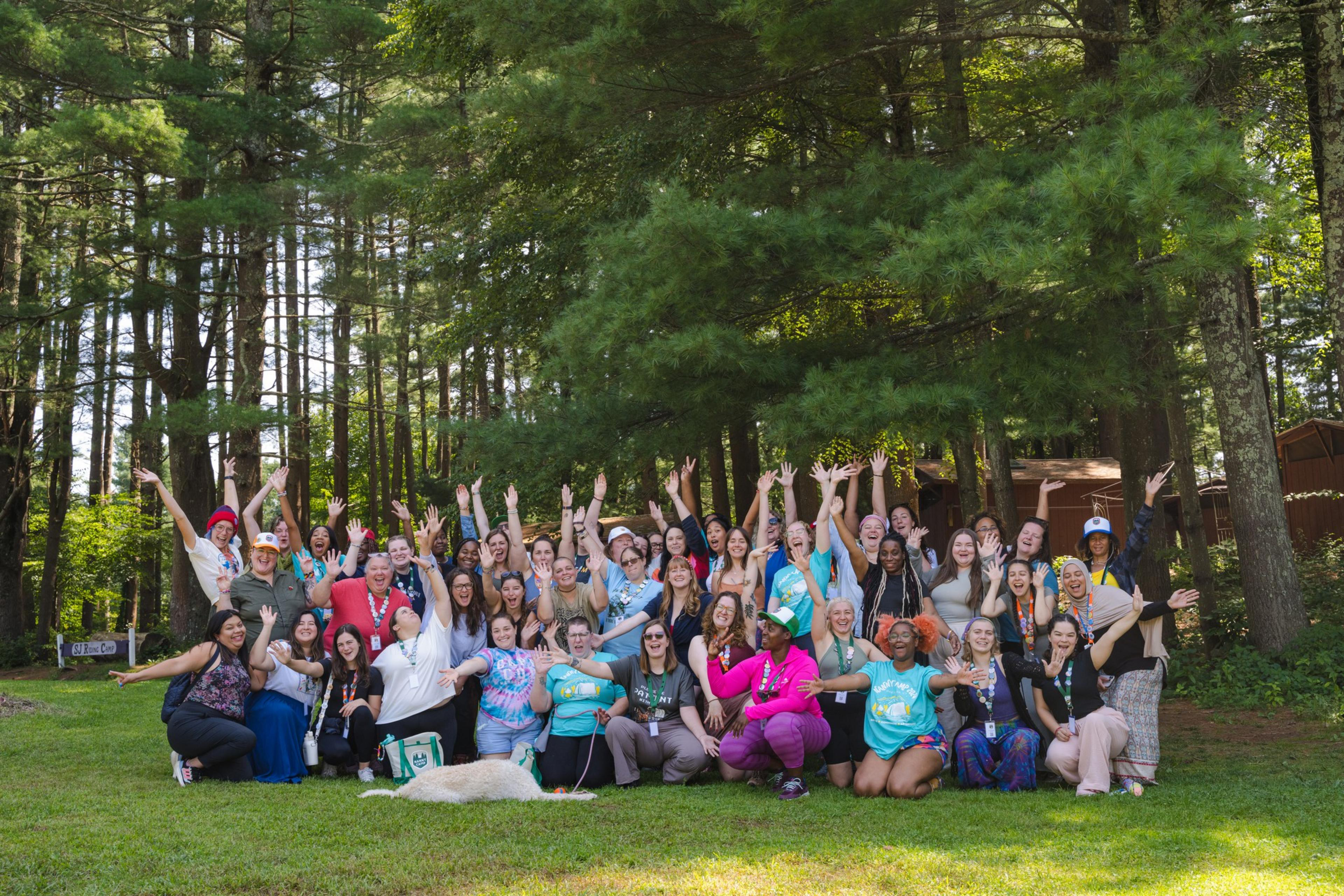 A large group of diverse people pose happily outdoors in front of tall trees, many with arms raised, while a white dog lies on the grass in front.