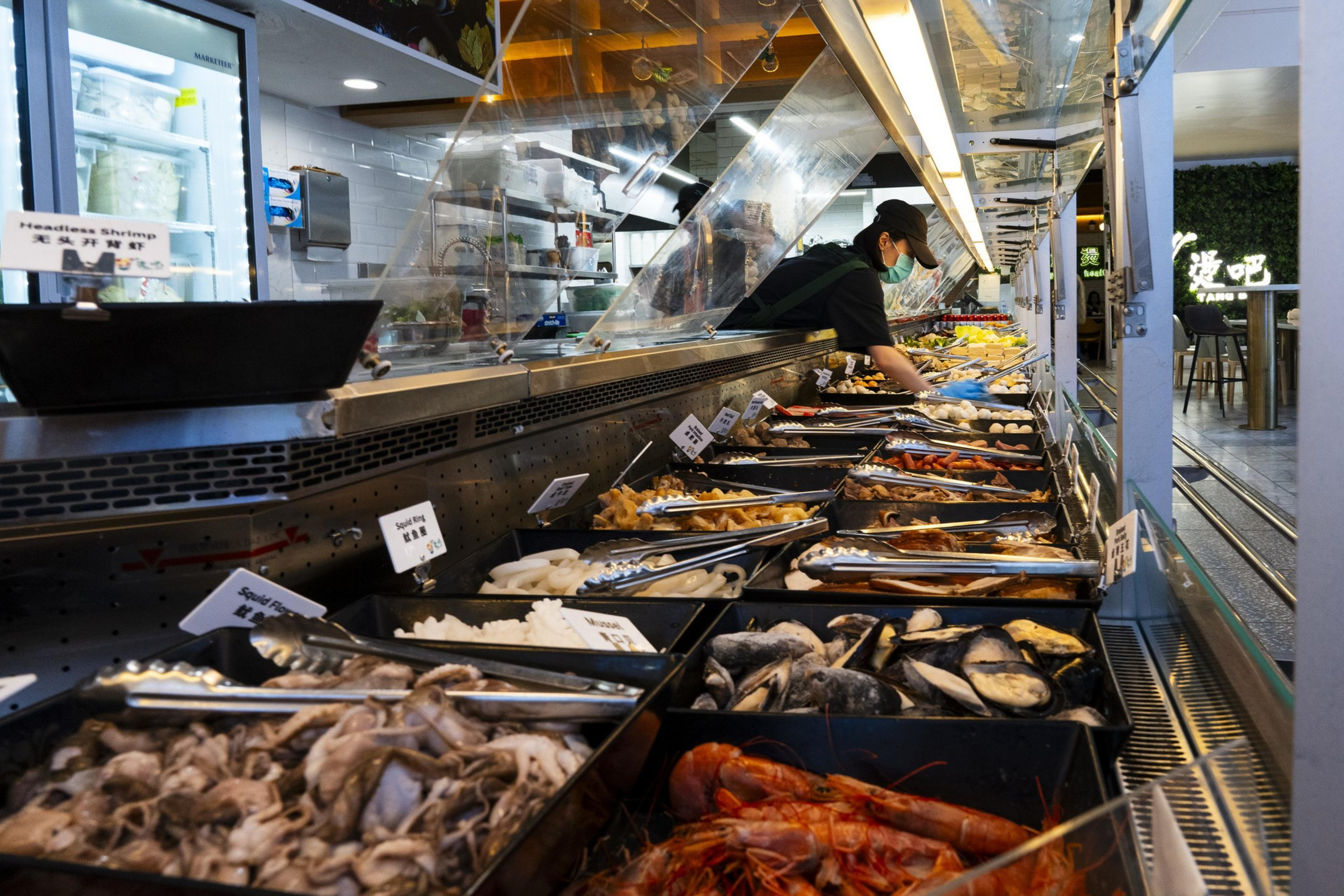 A person wearing a mask and cap arranges a diverse seafood buffet. The counter displays seafood like shrimp, squid, and mussels, each labeled with signs.