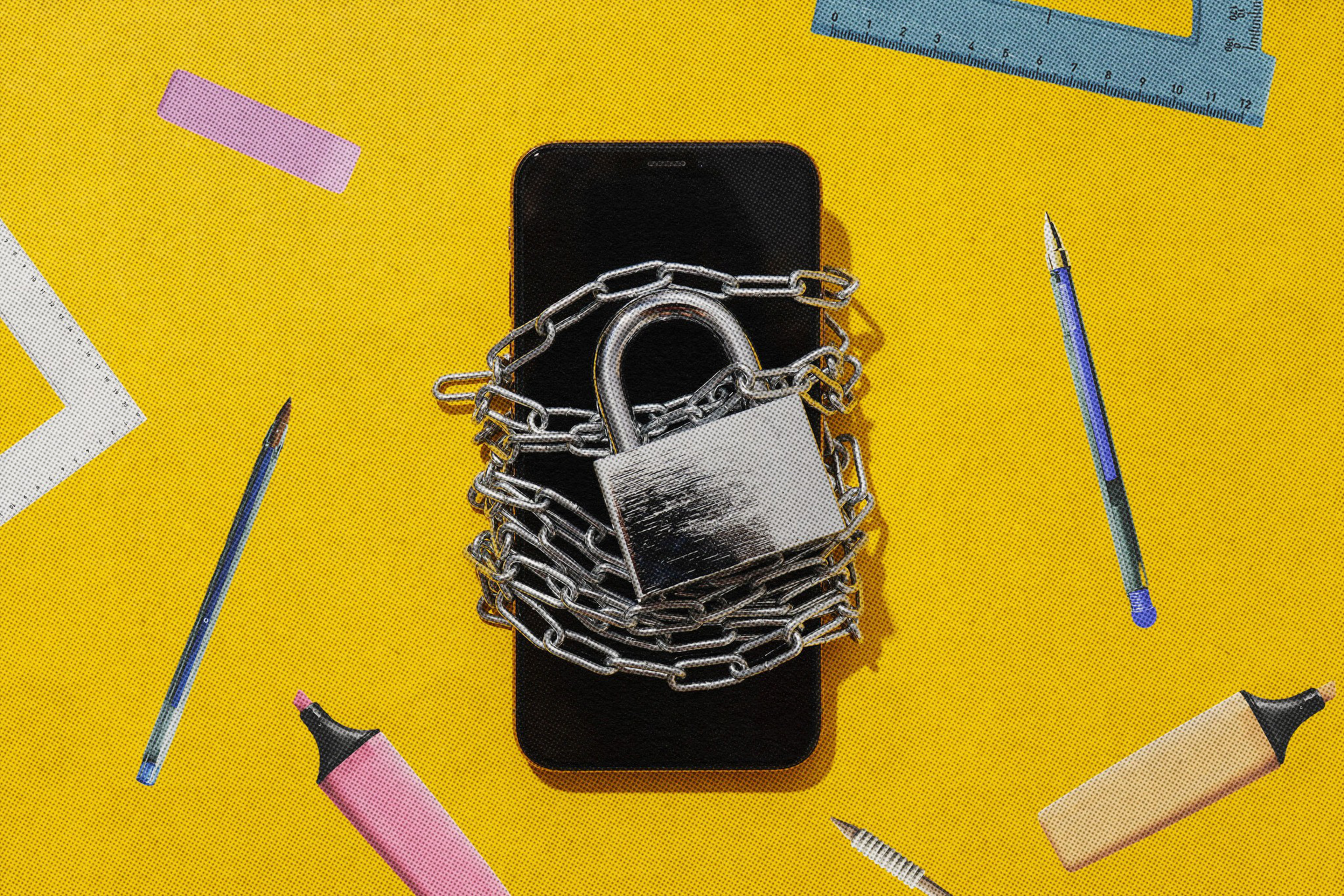 A smartphone wrapped in a chain with a padlock lies on a yellow surface surrounded by pens, highlighters, and rulers.