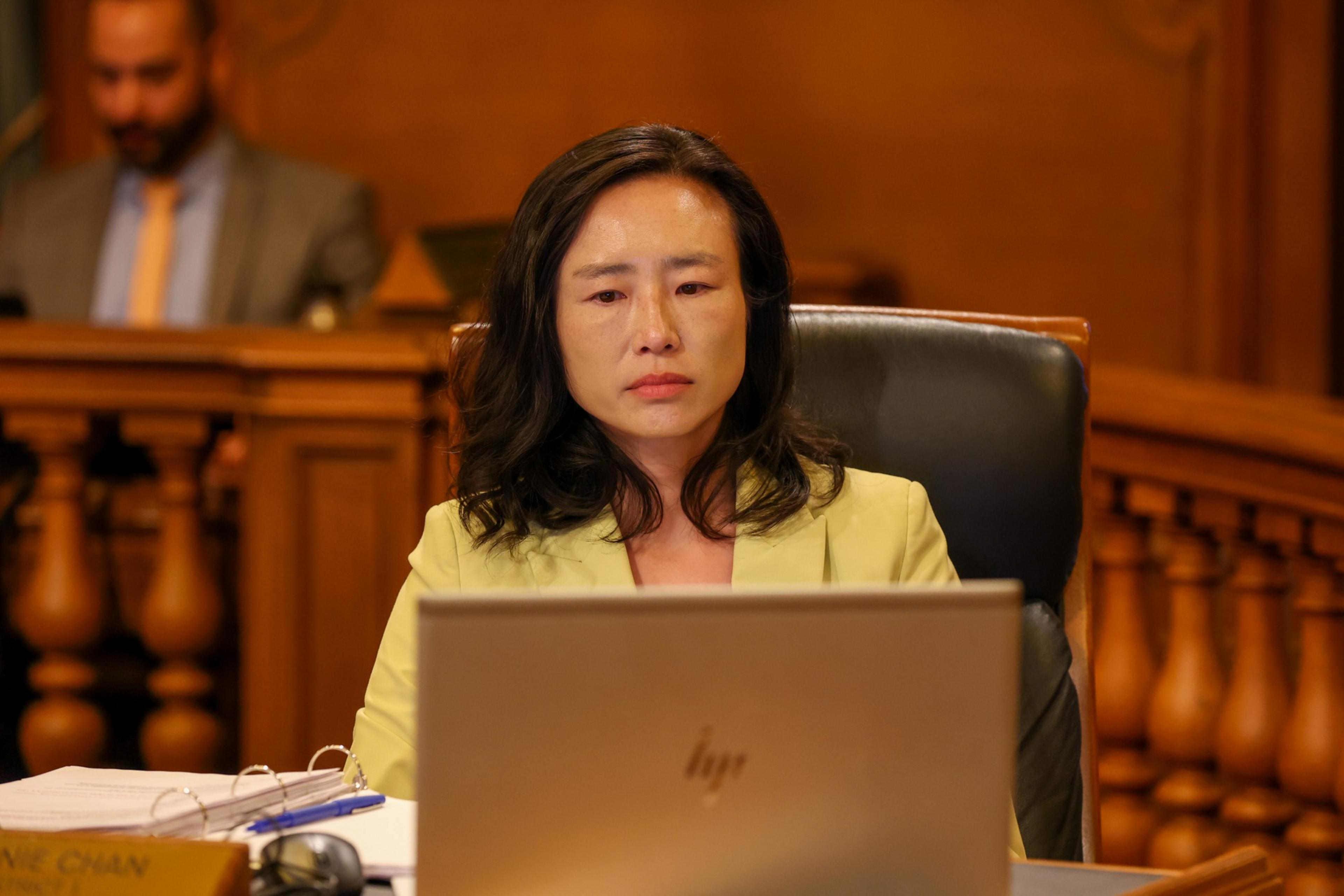 A woman in a yellow blazer sits seriously behind a laptop in a courtroom setting, with papers and a pen in front of her.