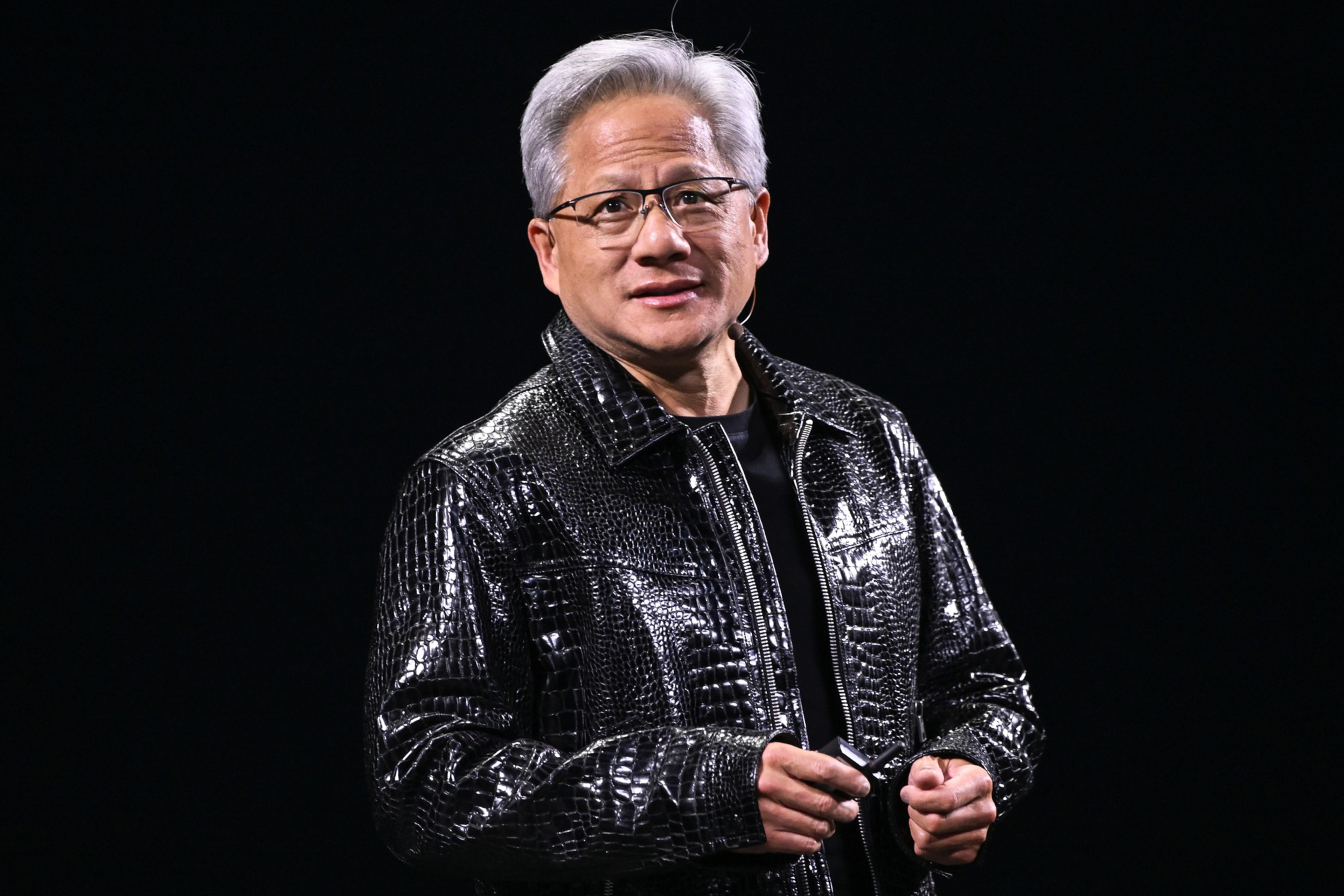 A person is wearing a shiny, black, textured jacket and glasses, standing against a dark background, holding a small object in their hands.