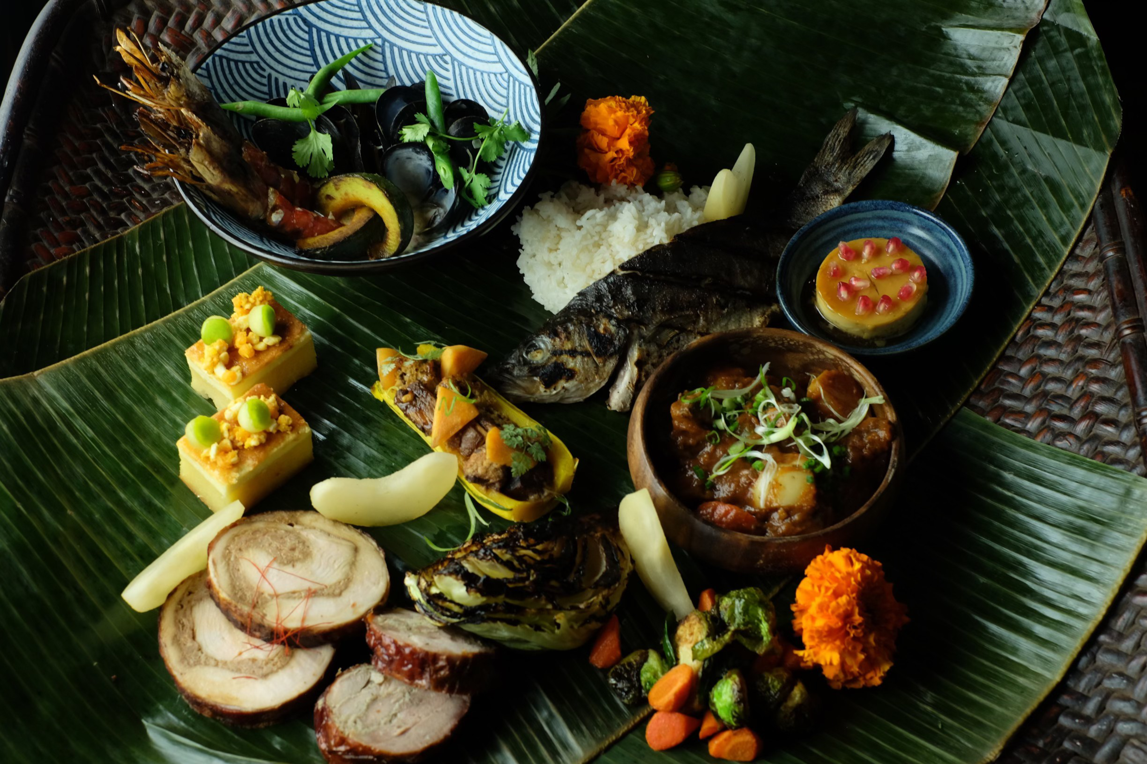 Grilled fish, rice, shrimp, vegetables, sliced meat, a stew, and small desserts are artfully arranged on large green leaves.