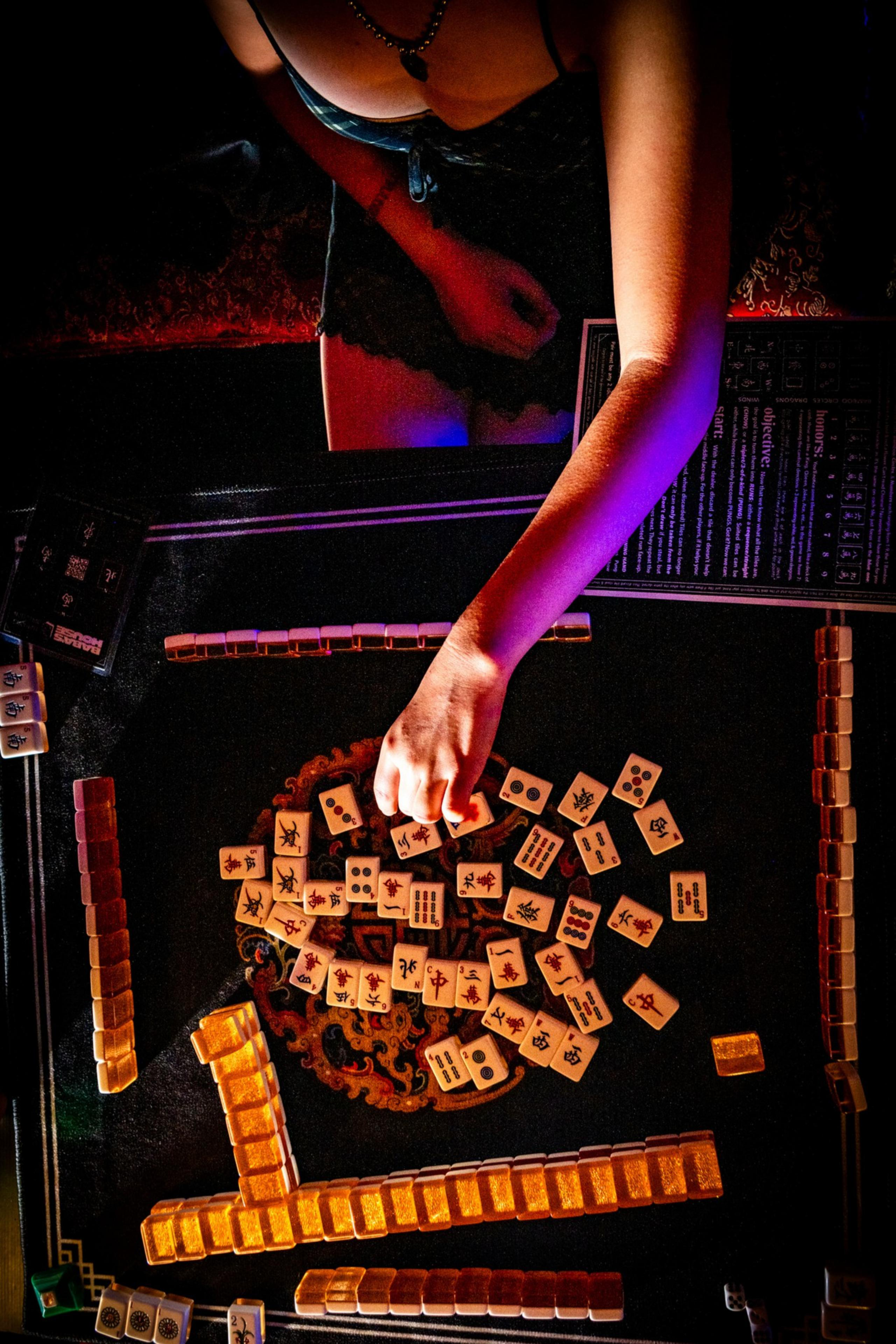 A person with a beaded necklace and a sleeveless top is reaching to pick a tile from a spread of Mahjong tiles on a dark surface with rows of face-down tiles surrounding the center.