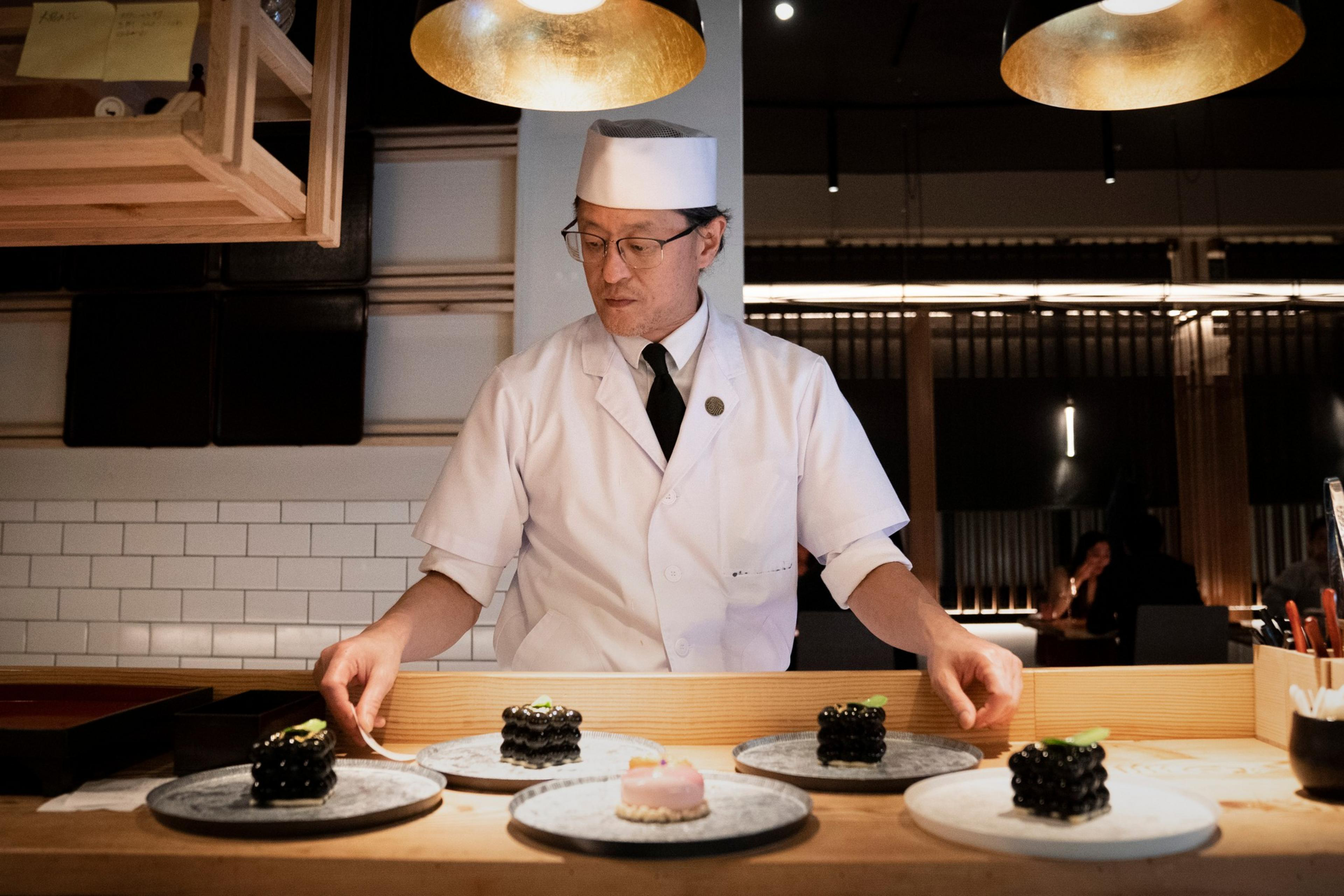 A chef in a white uniform and hat stands at a counter. He presents several intricately arranged dishes on plates, under warm lighting in a modern setting.