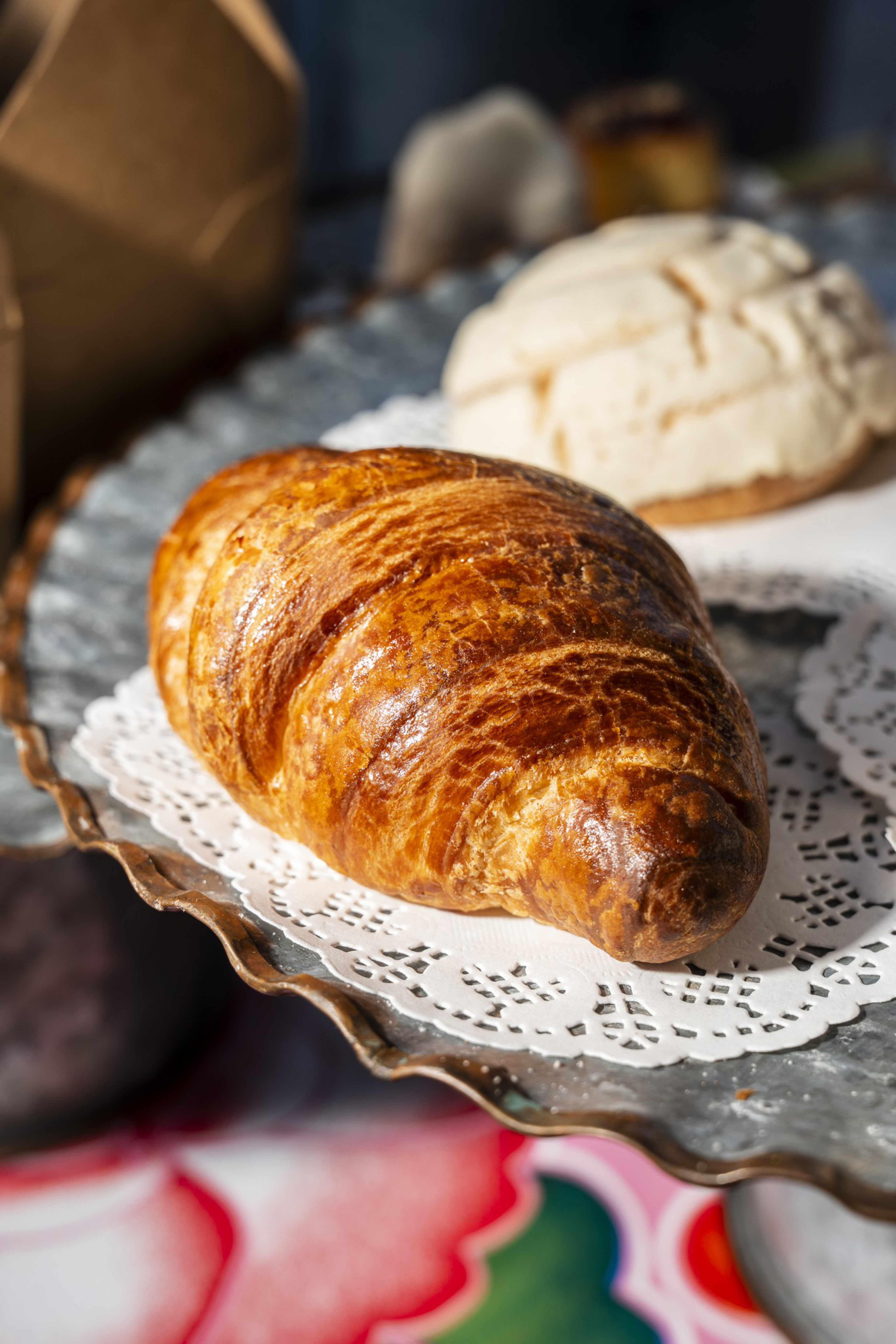 A golden-brown, flaky croissant rests on a lace doily atop a metal tray, with a lightly cracked round pastry in the background.
