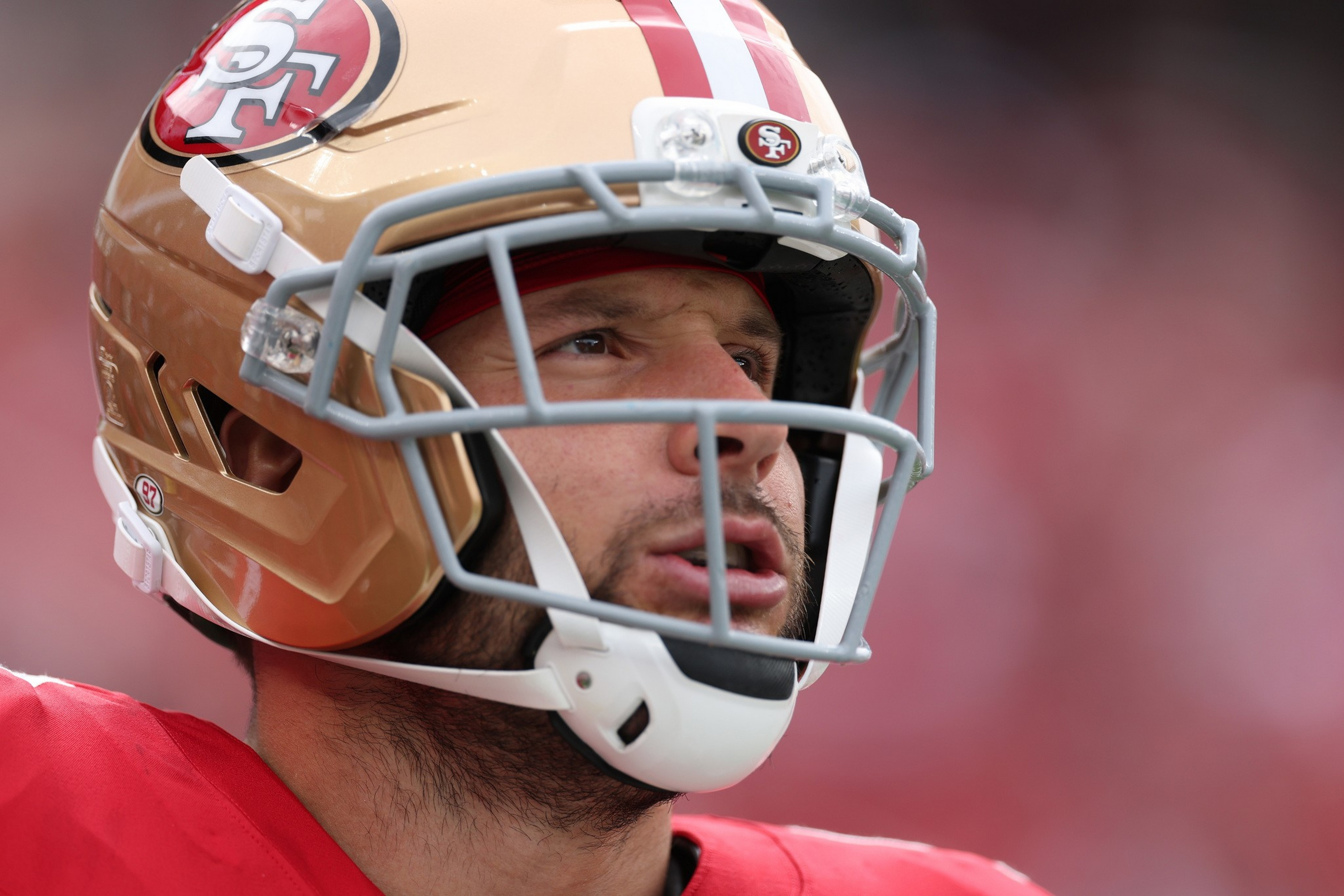 A football player wearing a gold helmet with a red and white stripe and a 49ers logo looks focused, with a red jersey visible.