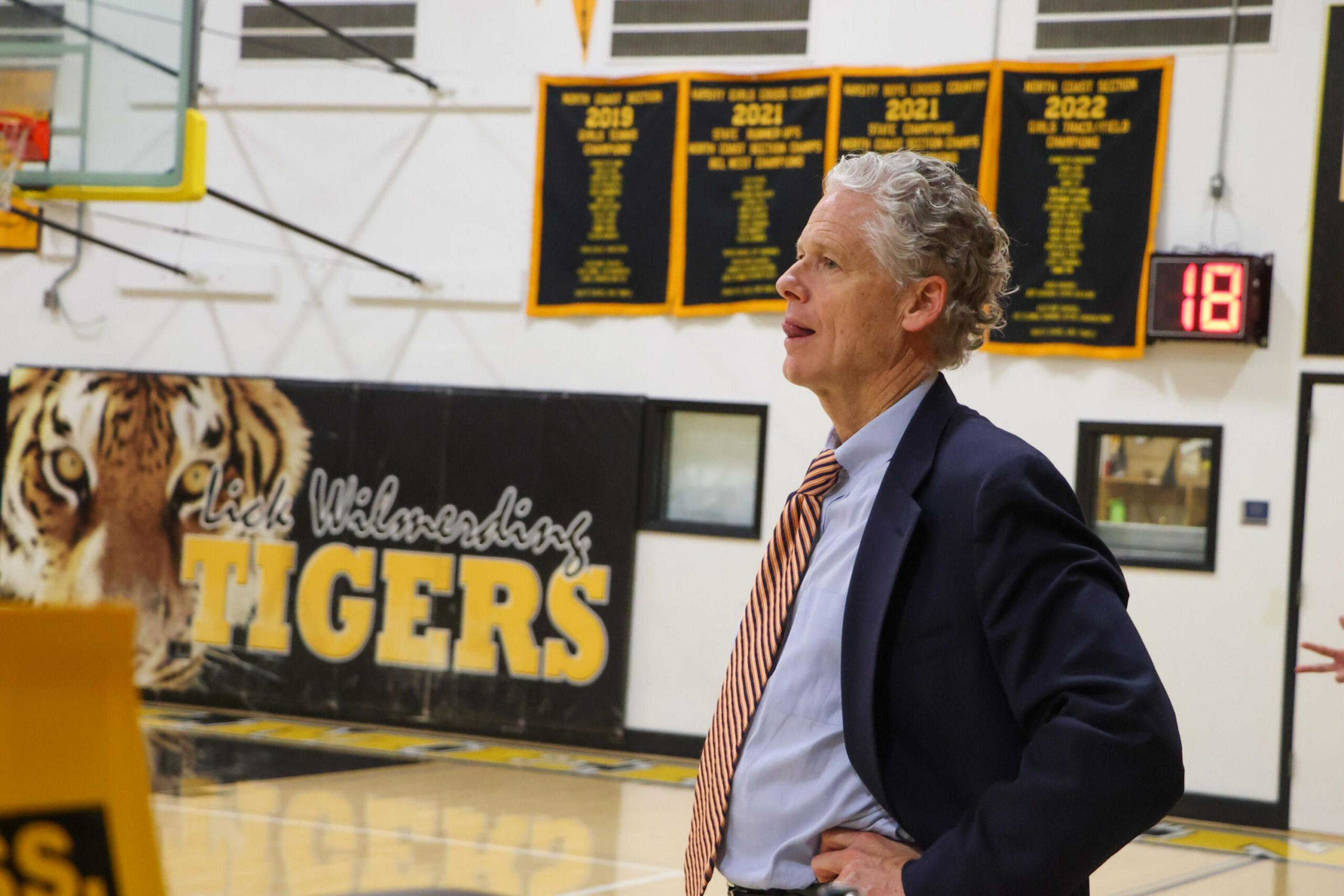 A man in a suit stands in a gym with his hands on his hips, looking to the side, with yellow and black school banners and a tiger mascot on the wall behind him.
