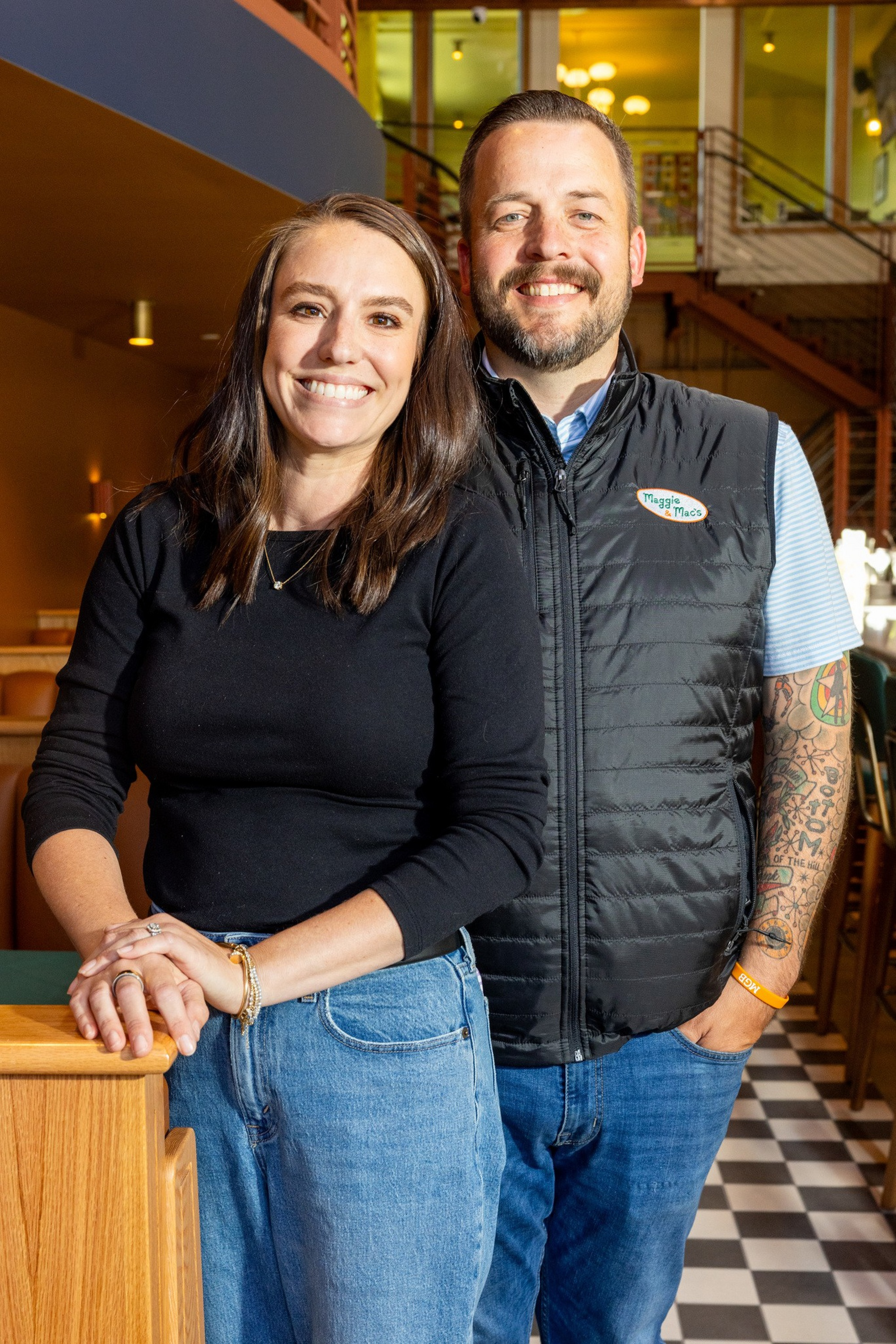 A smiling woman in a black top and jeans stands next to a smiling man wearing a black vest over a striped shirt with tattooed arms inside a cozy café.