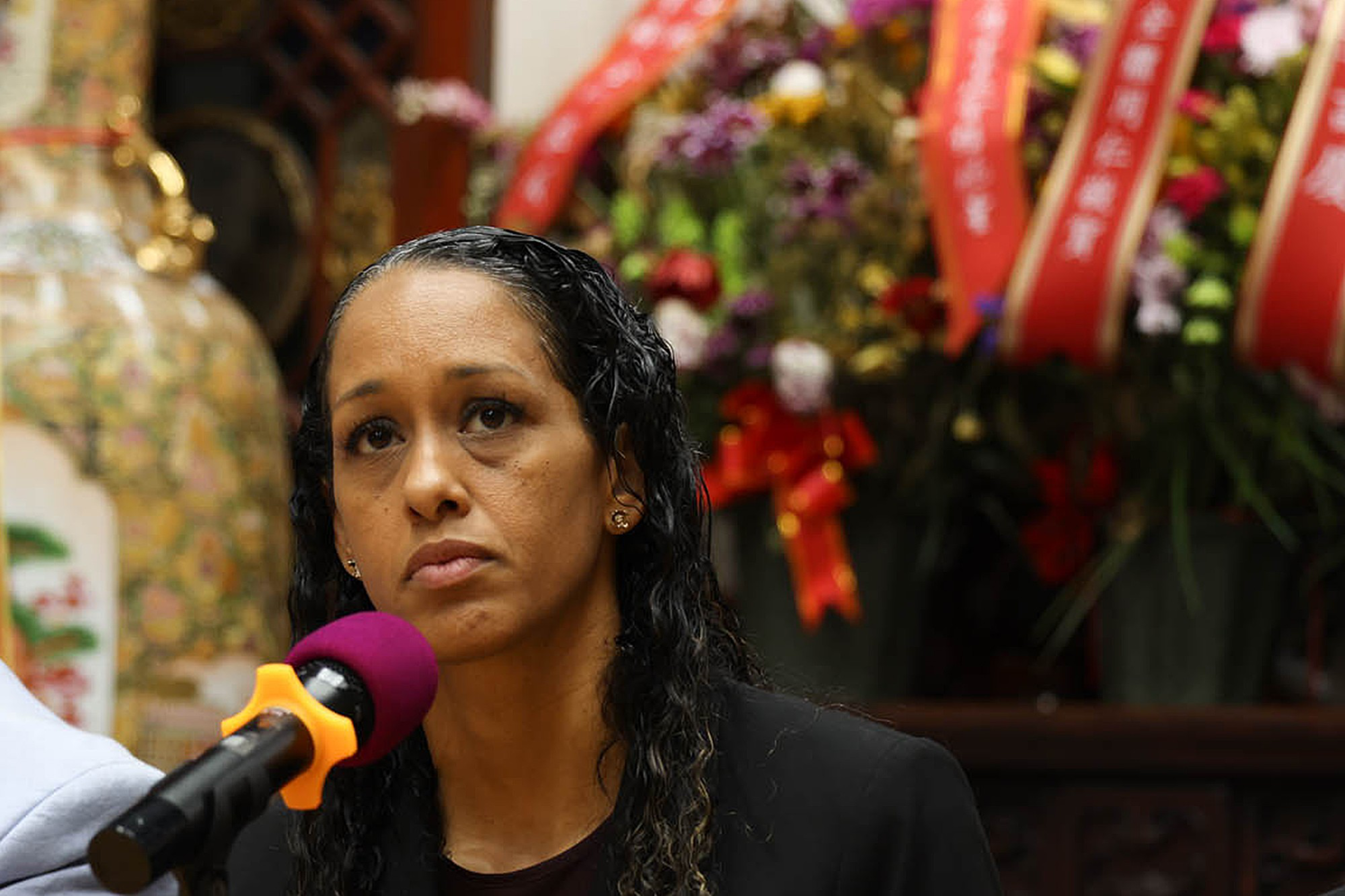 A serious woman with long curly hair speaks into a microphone, with a large floral arrangement and decorative vase in the background.