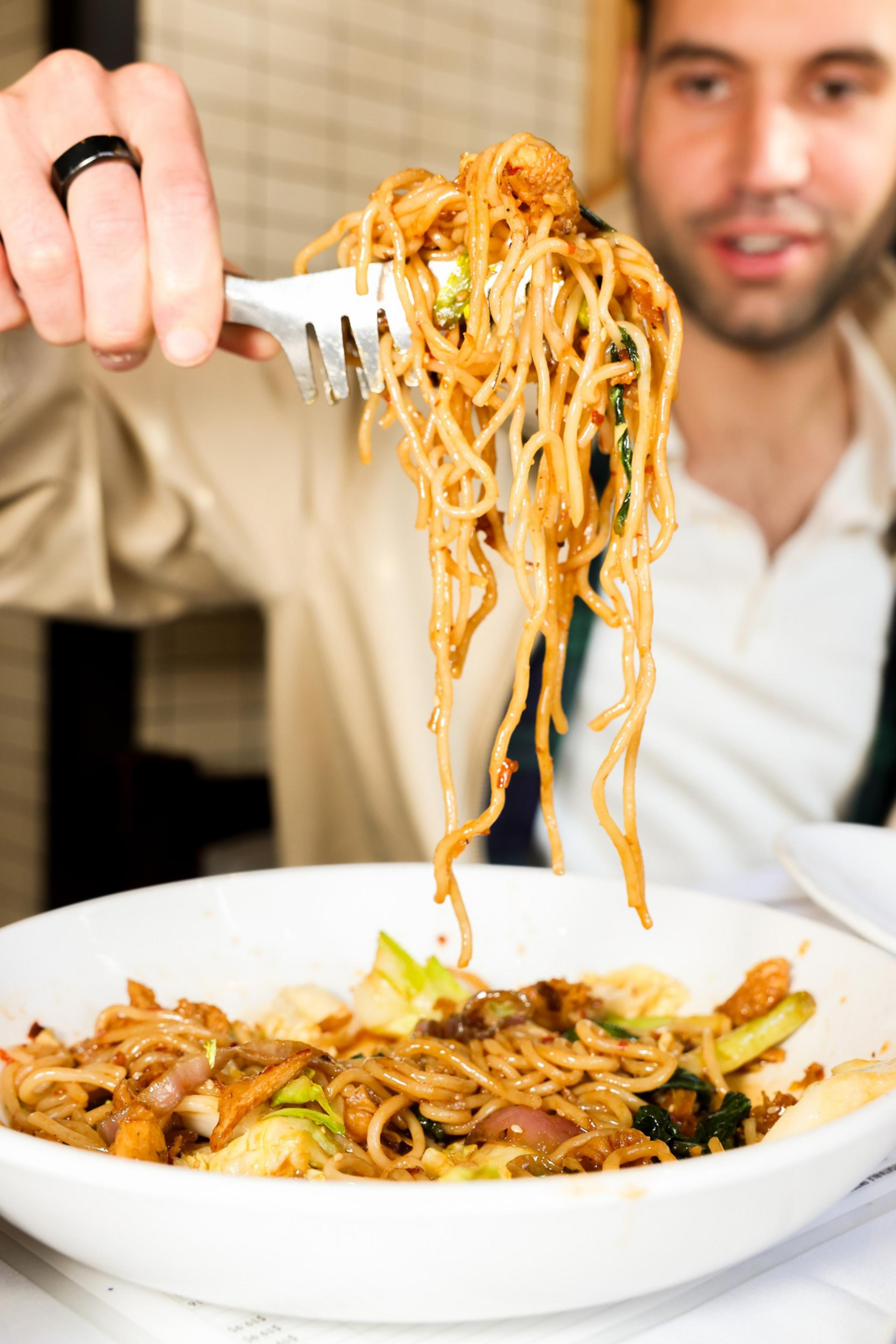 A person is lifting a forkful of stir-fried noodles from a large white bowl, with visible vegetables. The focus is on the noodles, hinting at enjoyment.