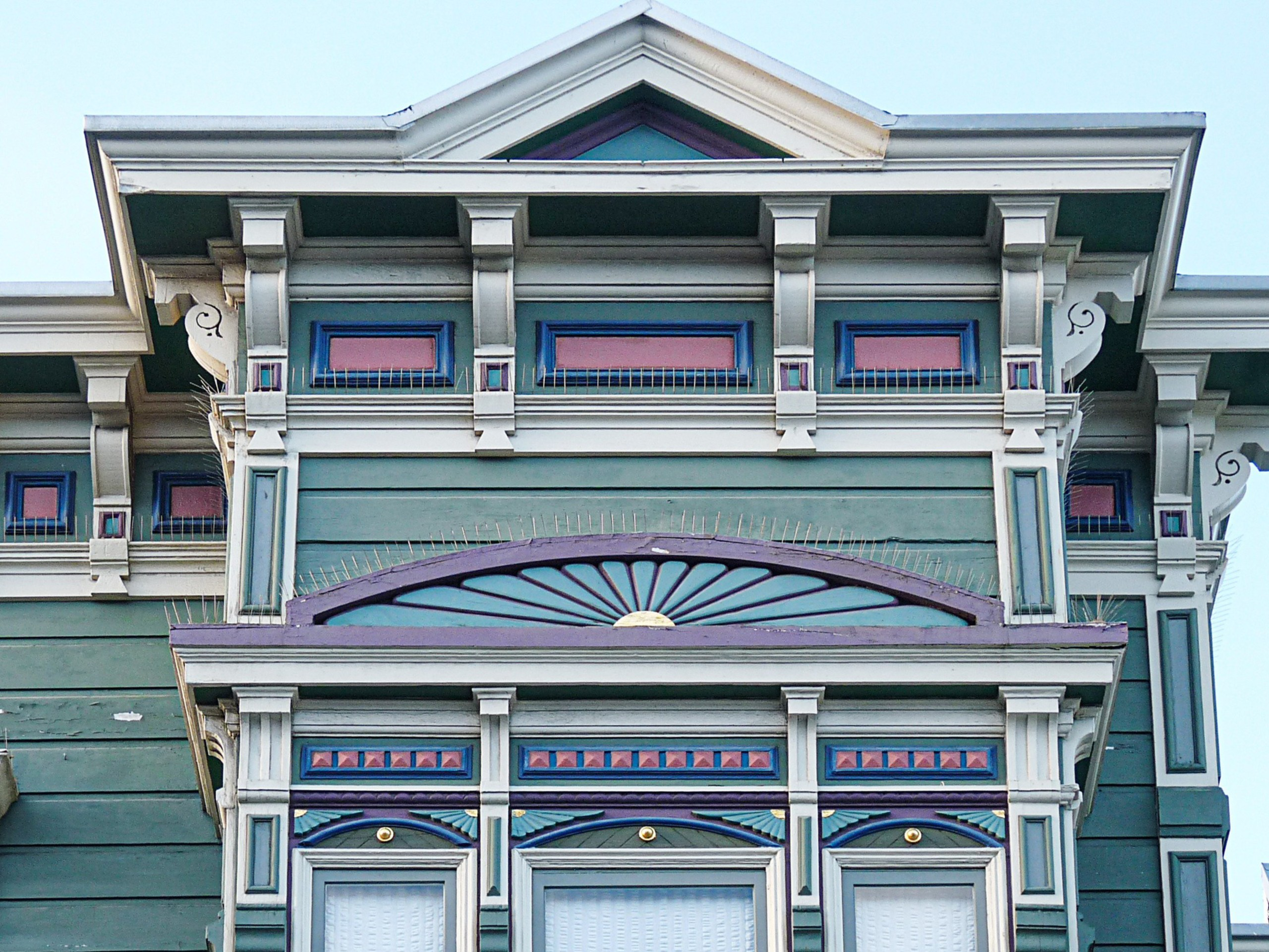 Green Victorian-style house with ornate white trim, purple and blue accents, small rectangular windows, and decorative architectural details near the roof.