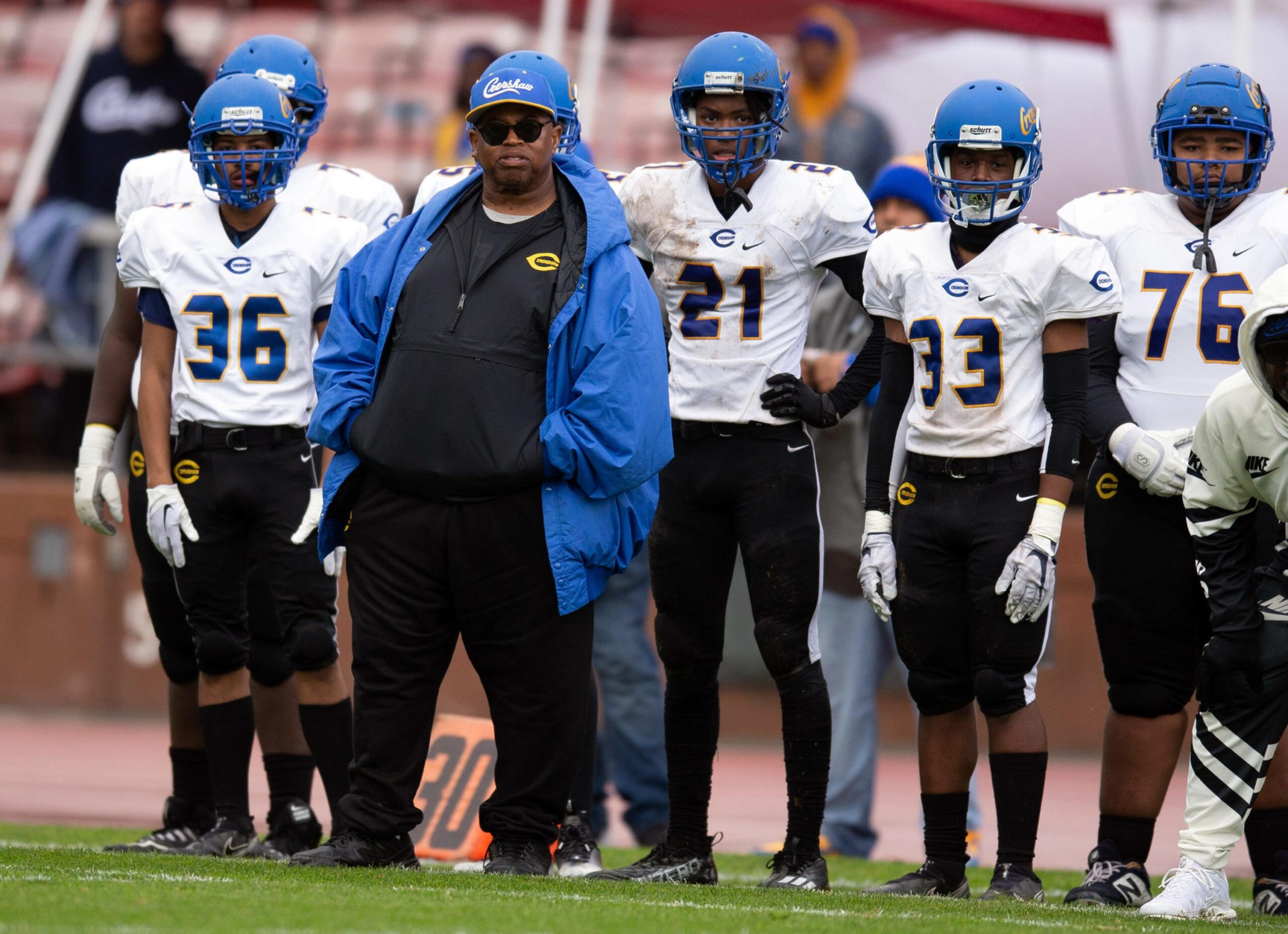 A football coach in a blue jacket and cap stands with five players in white jerseys and blue helmets on the sidelines of a football field.