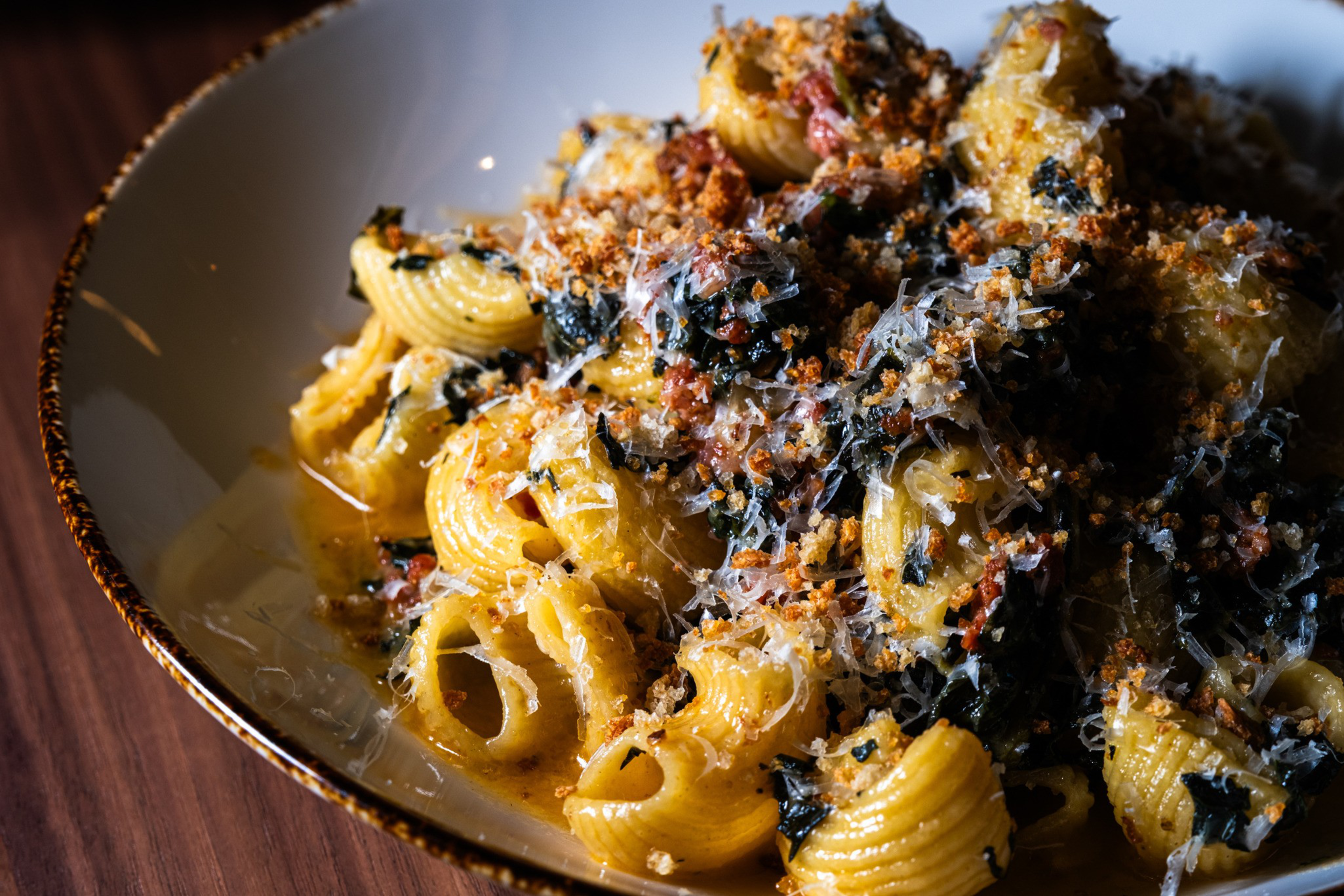 A bowl of pasta with a rich, creamy sauce is topped with crispy breadcrumbs, finely grated cheese, and pieces of dark leafy greens.