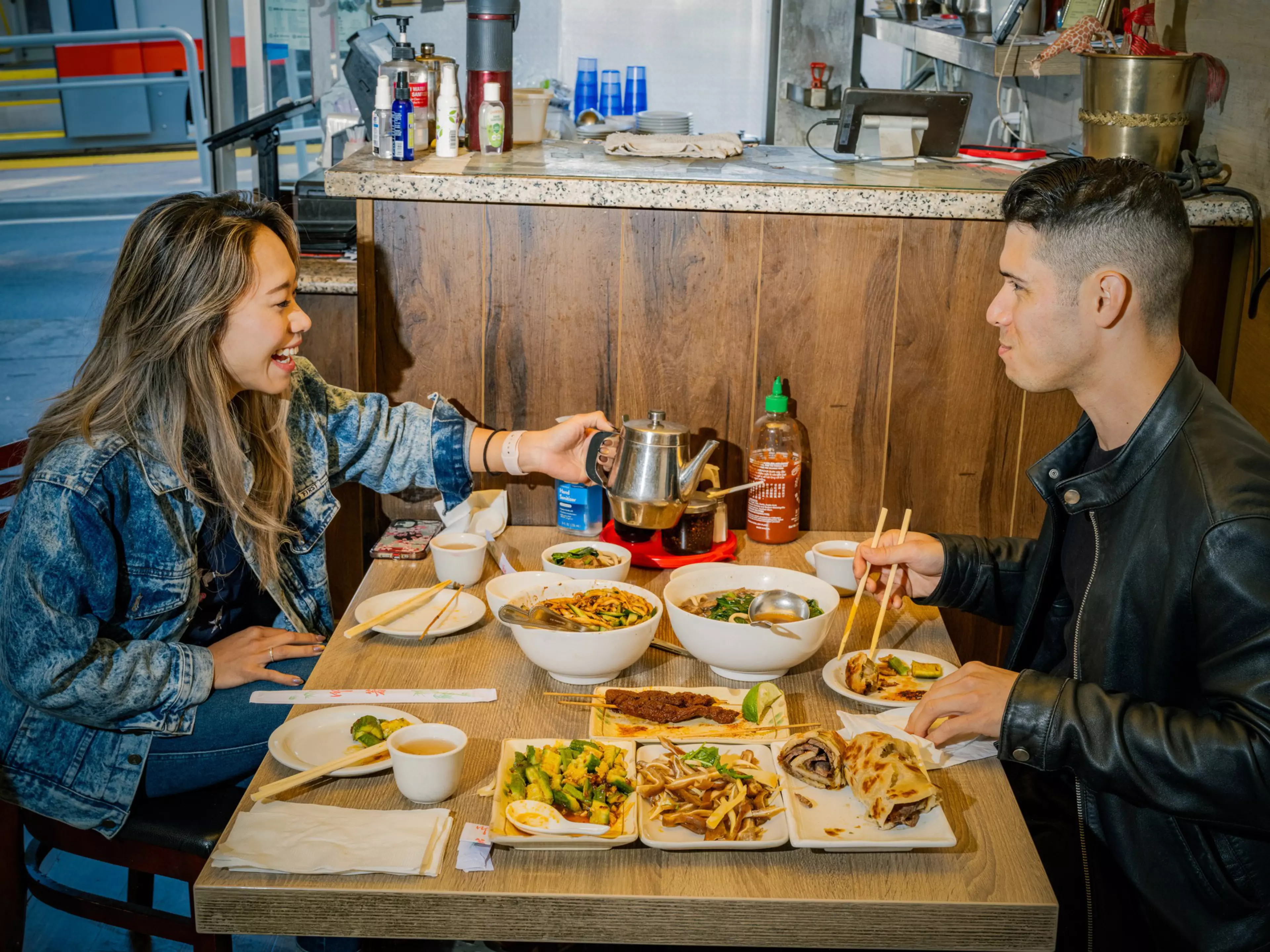 A woman and a man share a meal with various Asian dishes on a wooden table, using chopsticks and a metal teapot in a casual dining setting.
