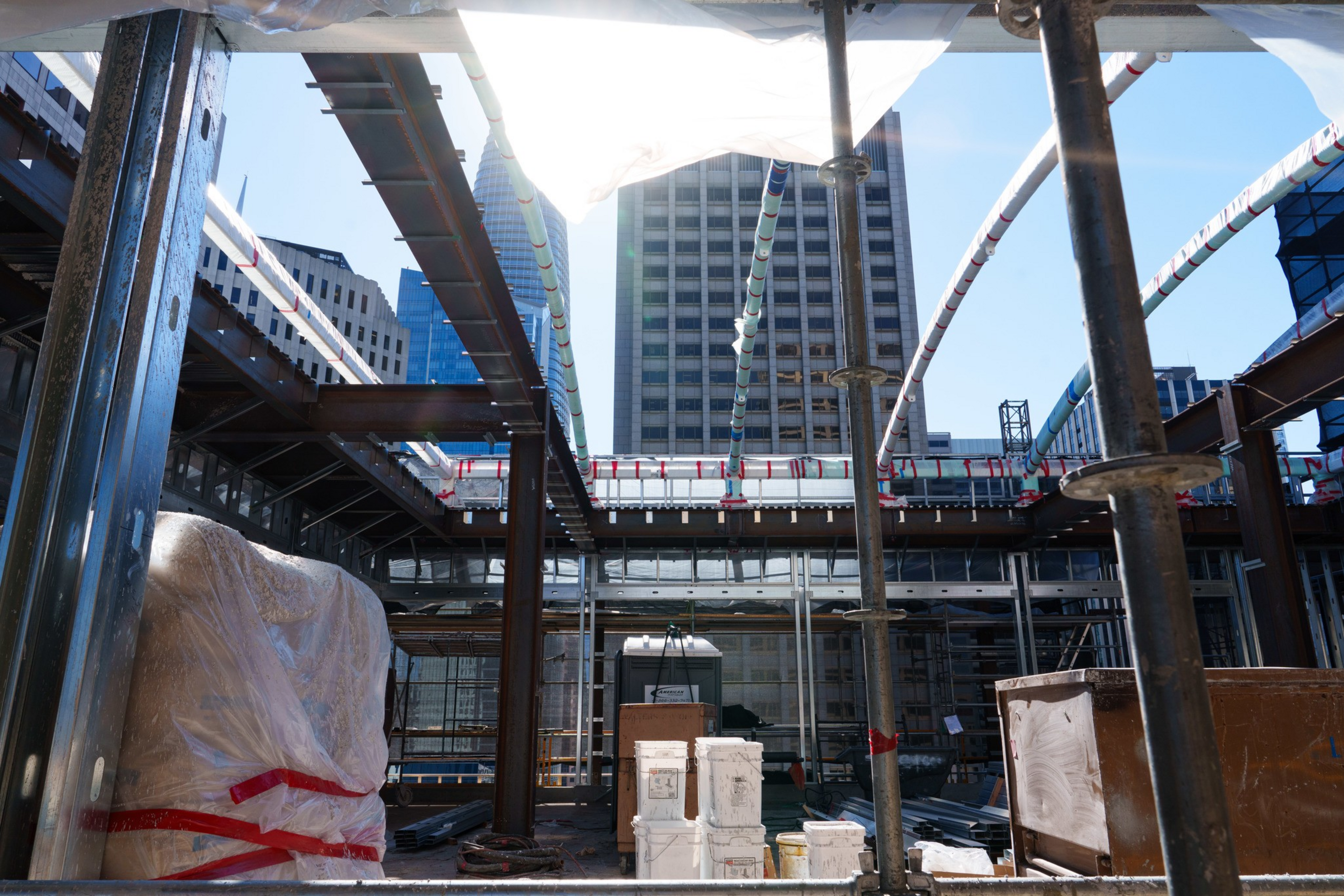 Steel beams and construction materials are arranged on a building site with tall urban buildings visible in the background under a clear blue sky.