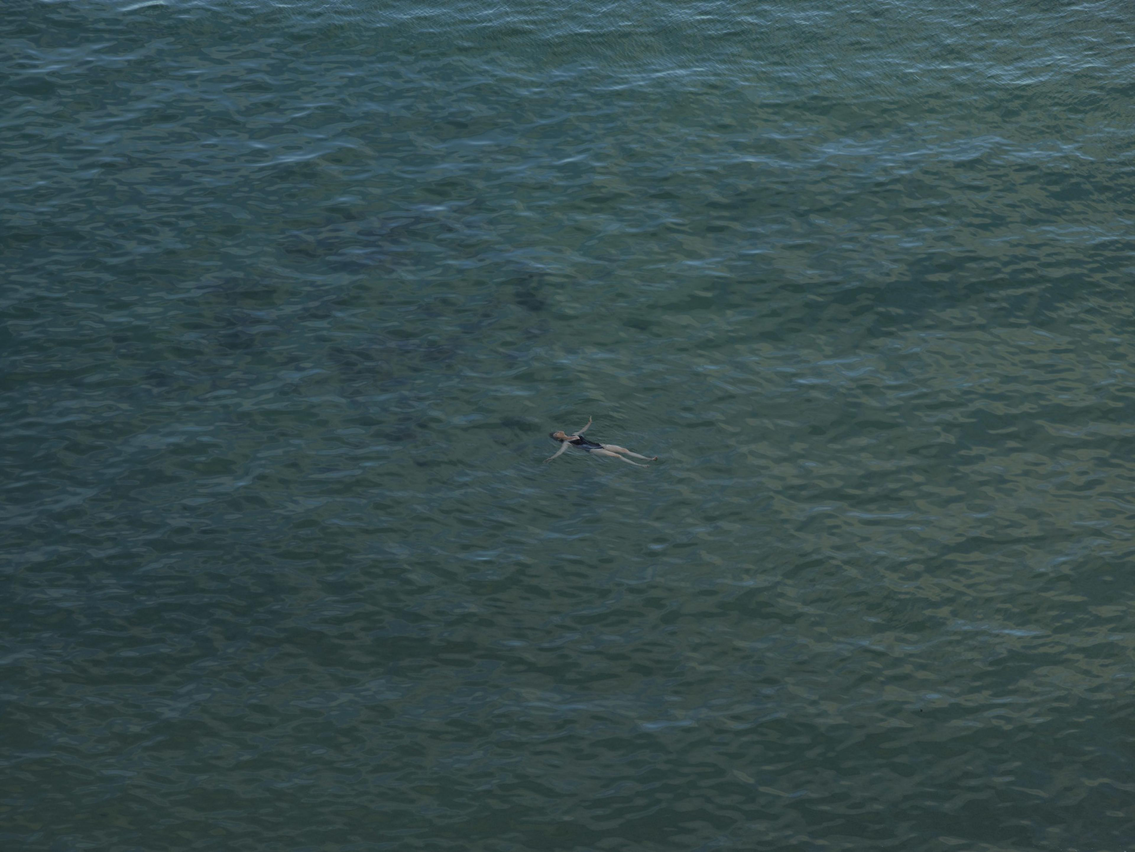 A person is floating in clear, calm blue-green water, surrounded by gentle waves. The scene appears serene and peaceful.