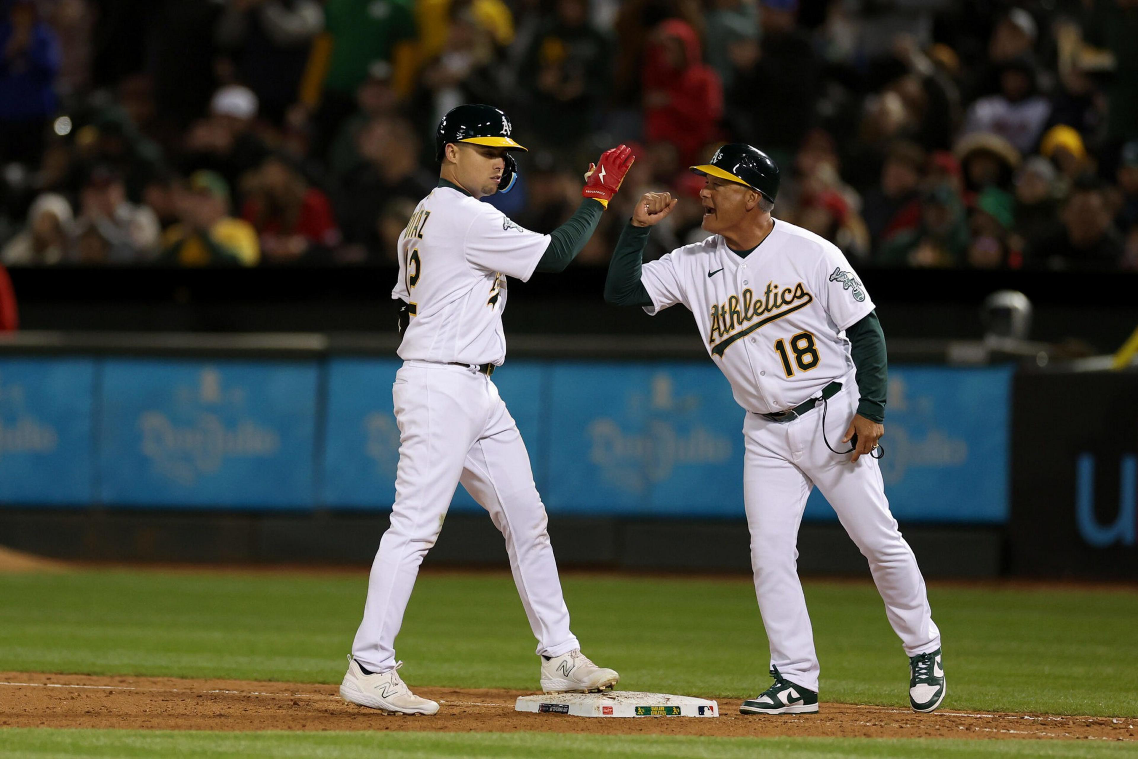 Two baseball players from the Athletics team celebrate at a base. The player in a white uniform and helmet appears to have reached the base safely, as the coach, also in white, celebrates.