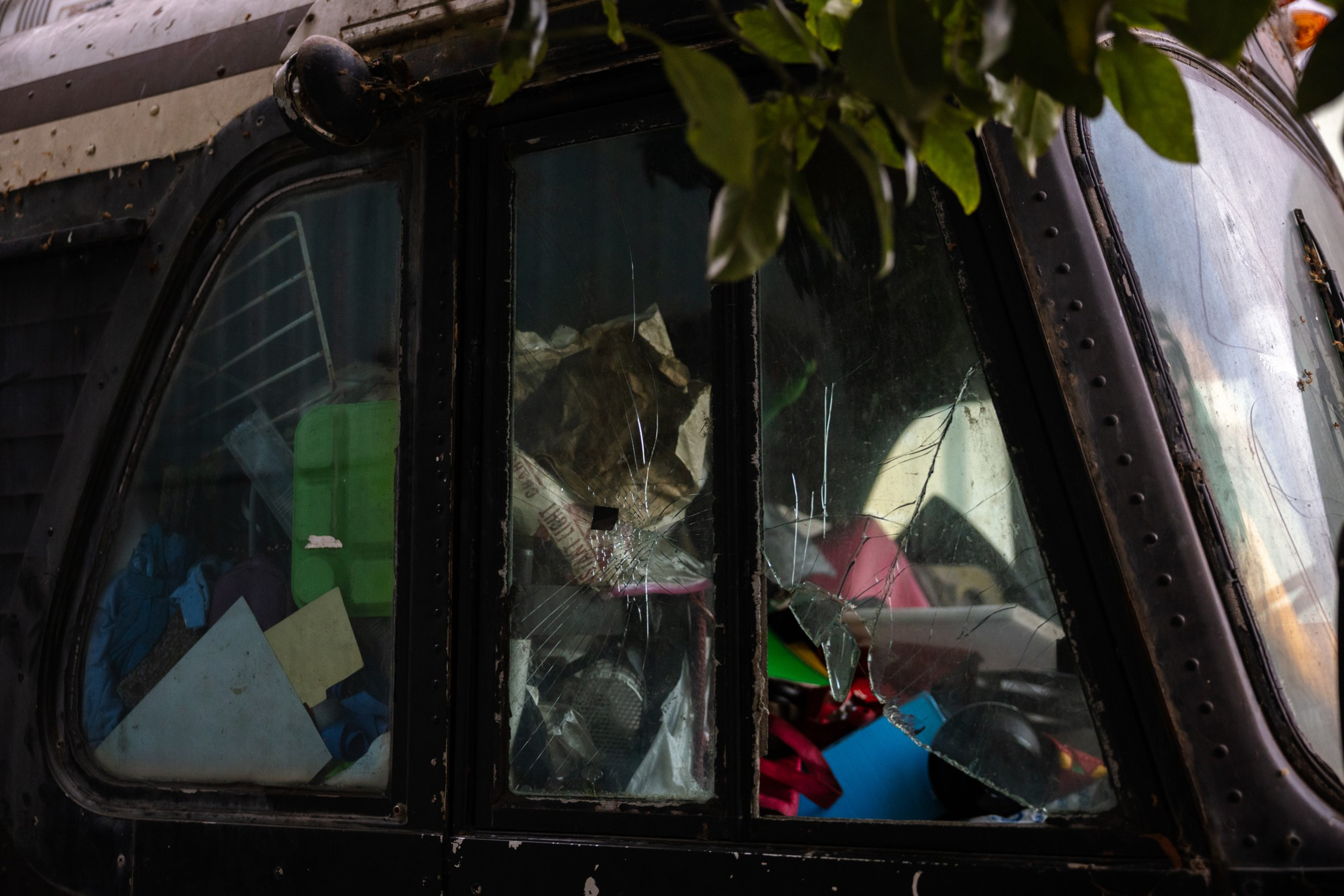 A shattered window on a dark vehicle reveals cluttered, colorful objects inside, partially obscured by leaves hanging above.