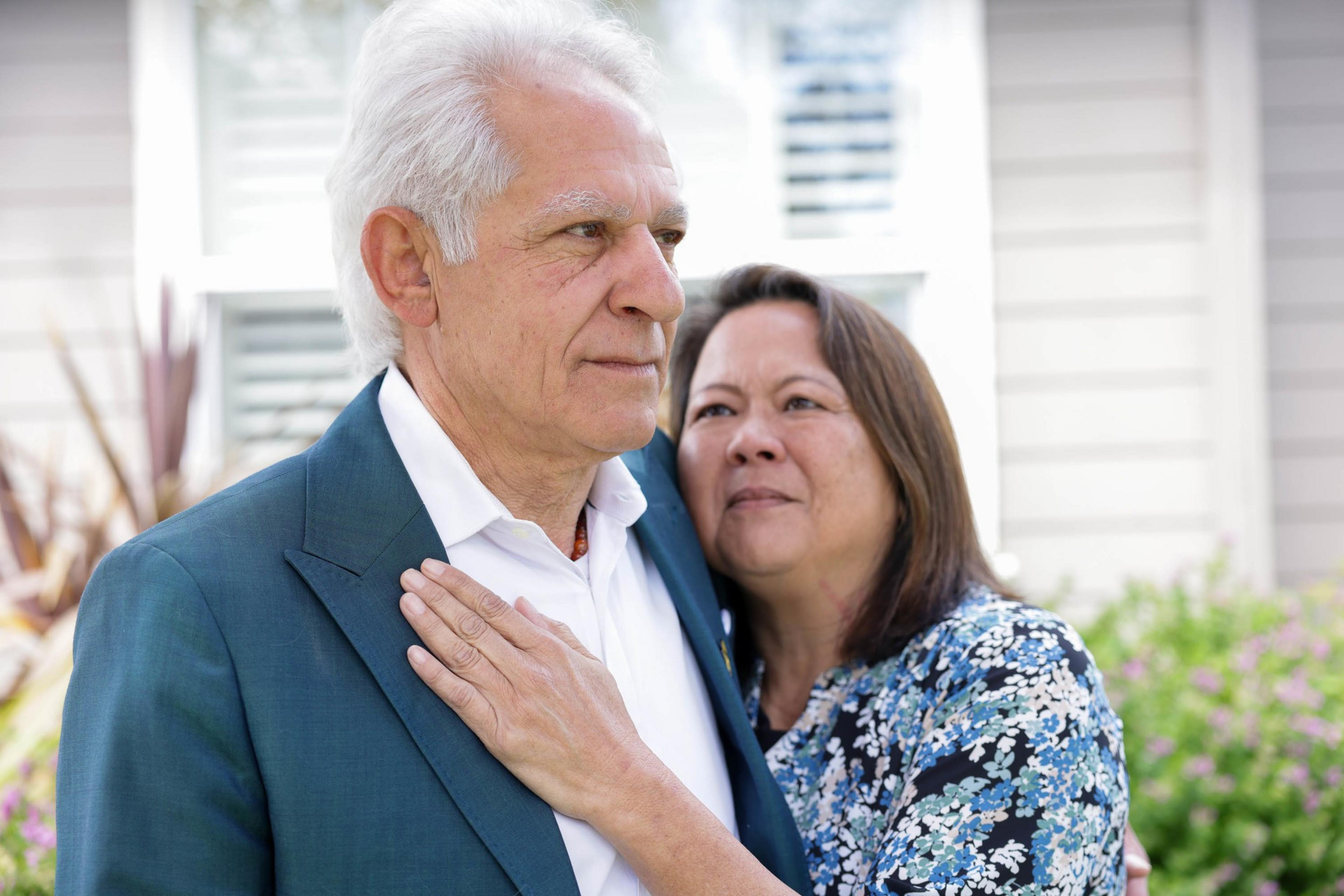 An older couple stands close, the woman embracing the man from behind, both looking thoughtful, in daylight. 