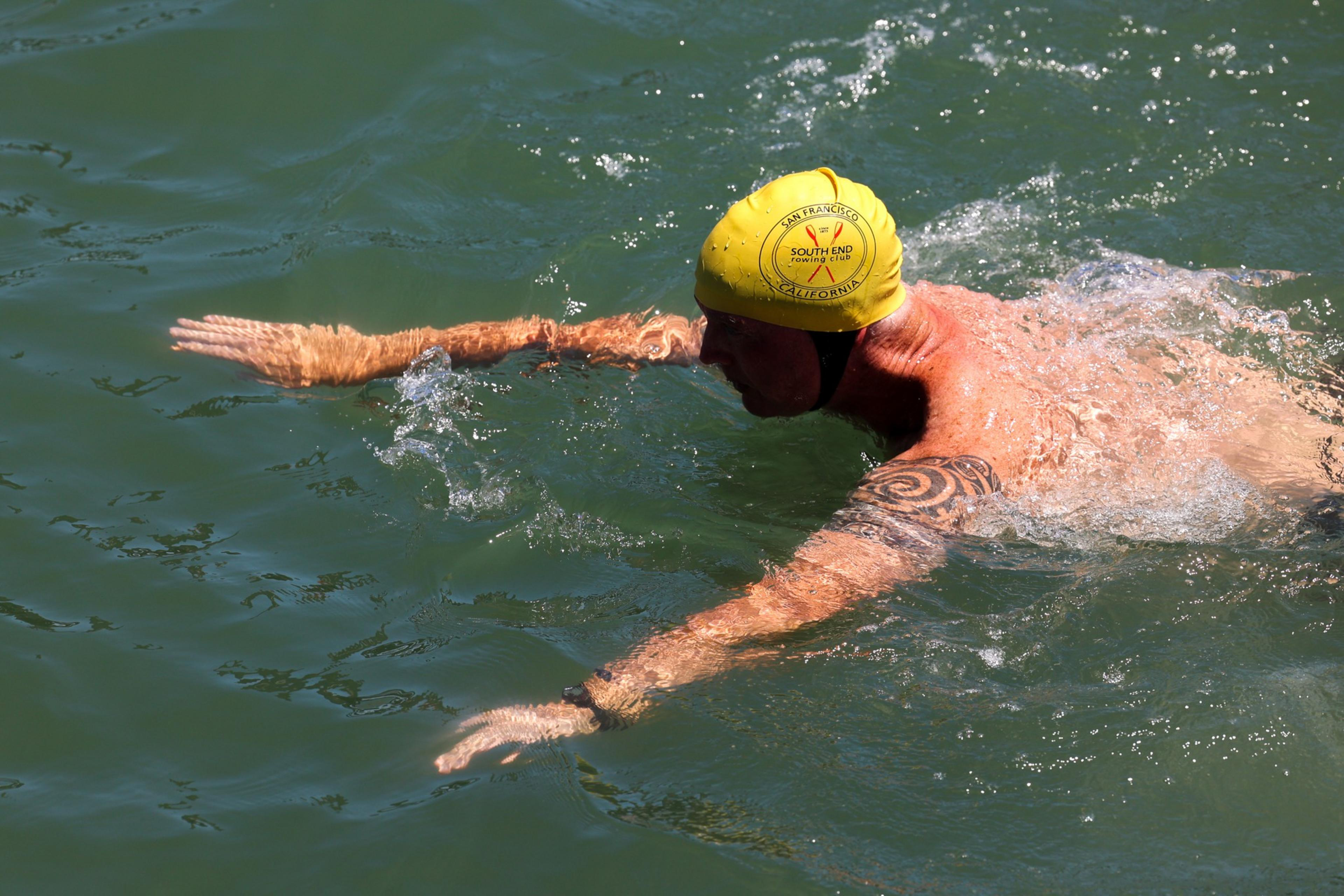 A swimmer wearing a yellow swim cap with San Francisco South End Rowing Club logo is swimming freestyle in green water, showing a tattoo on their shoulder.