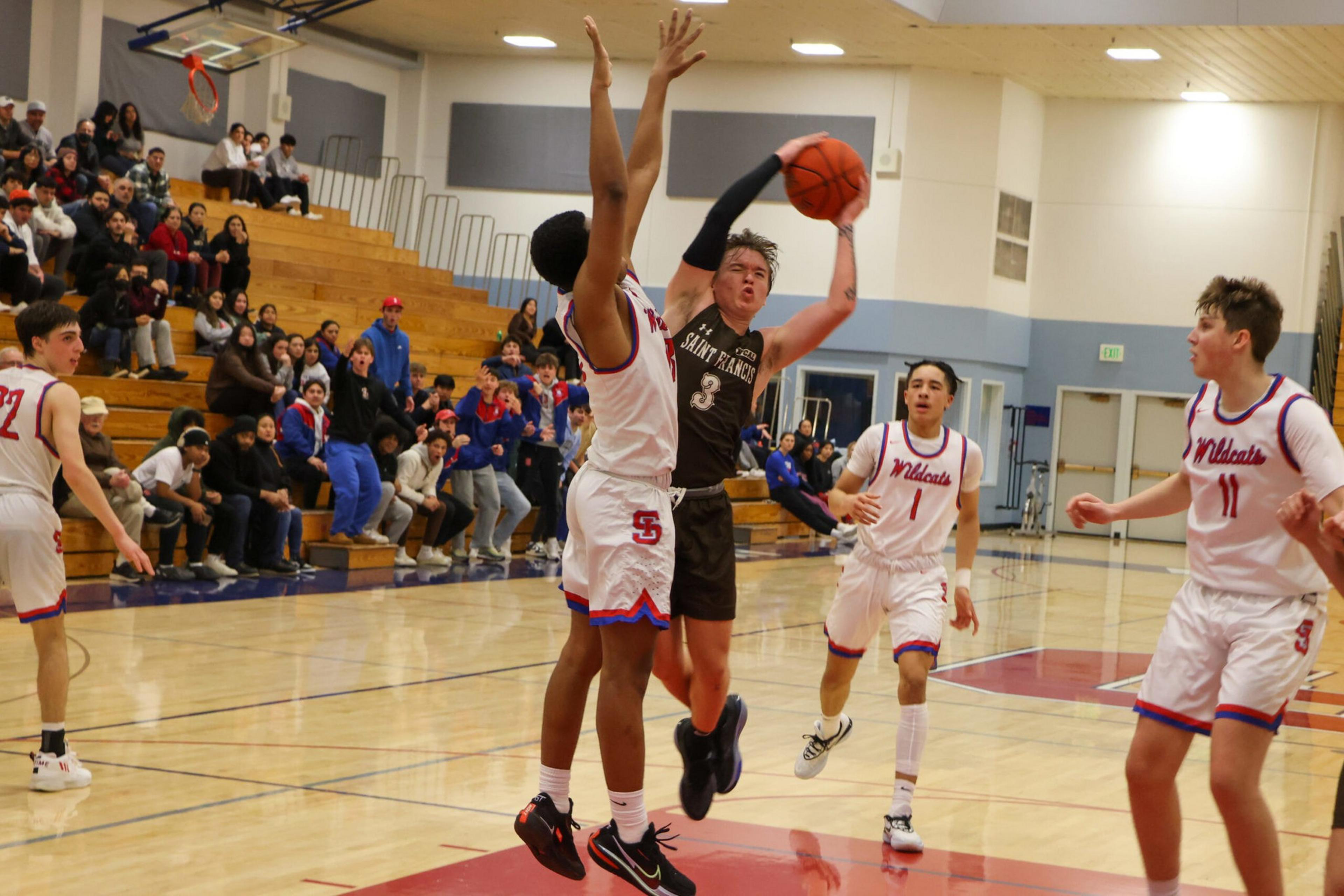 A basketball player in a black uniform jumps to shoot while three defenders in white uniforms with "Wildcats" try to block him in a gym with spectators.