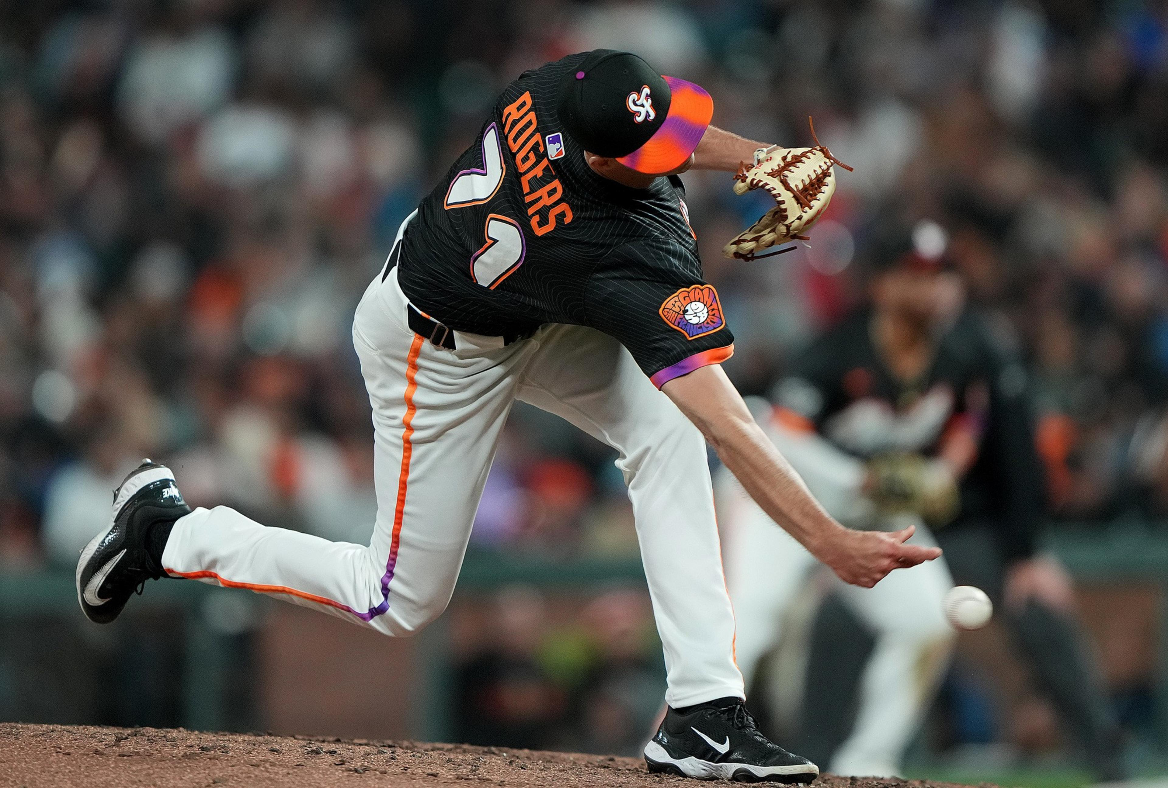 A baseball pitcher wearing a black jersey with "ROGERS" and number 71 is mid-throw on the mound, with a blurred player and crowd in the background.