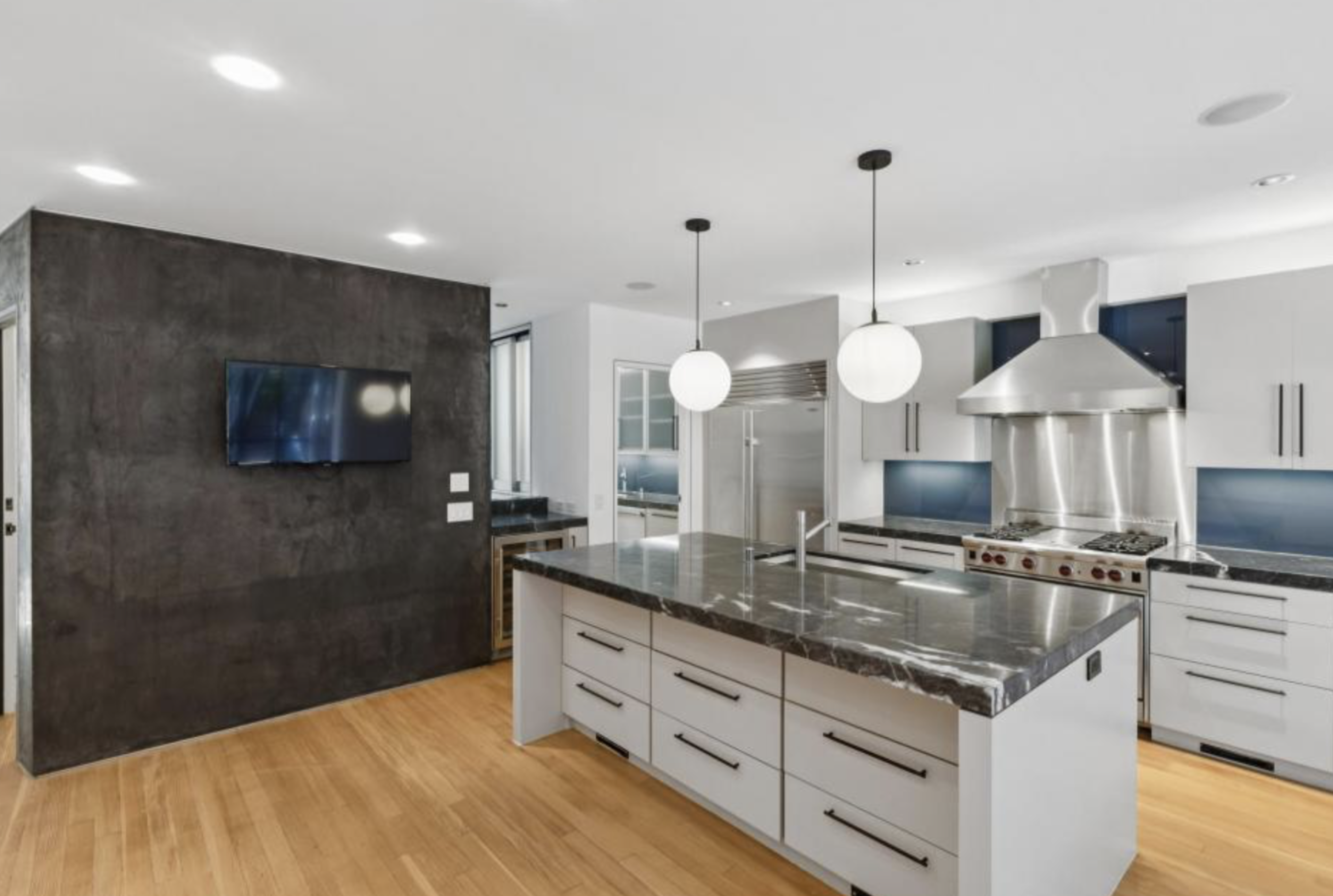 A modern kitchen with a large marble island, sleek white cabinets, stainless steel appliances, two round pendant lights, and a small wall-mounted TV.