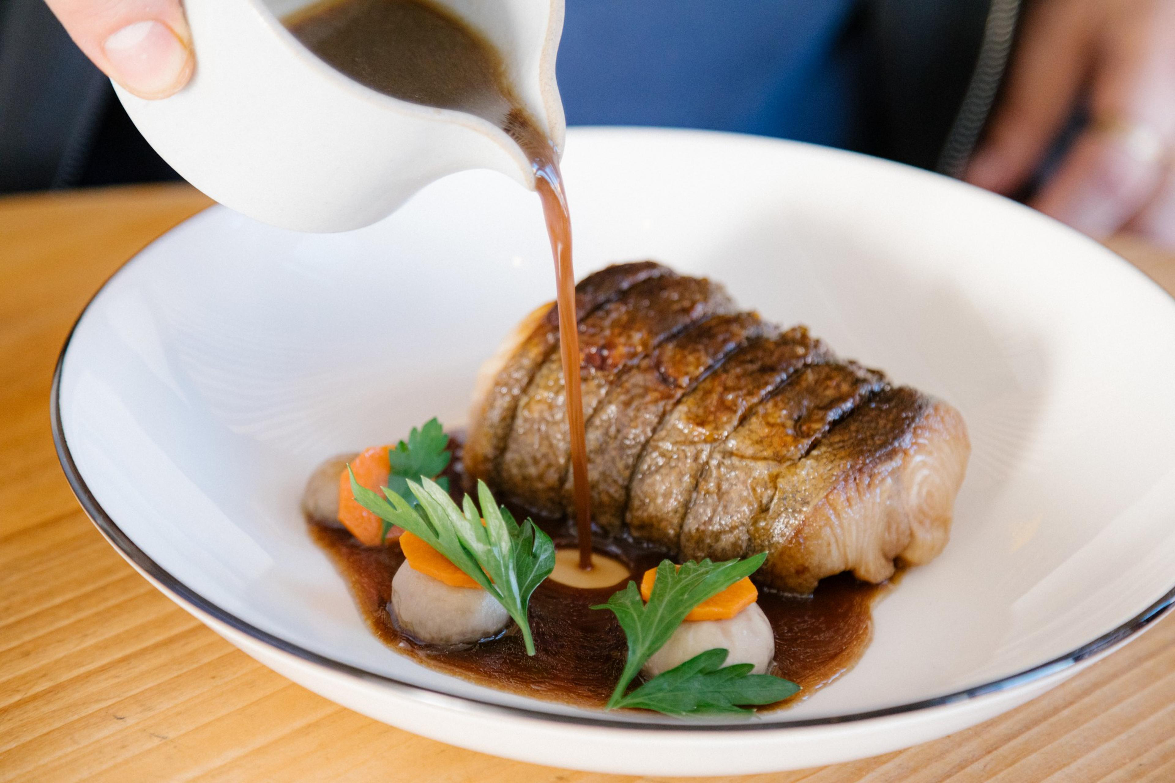 A hand pours sauce over a sliced, cooked piece of meat on a white plate. The dish is garnished with herbs and small vegetable slices.