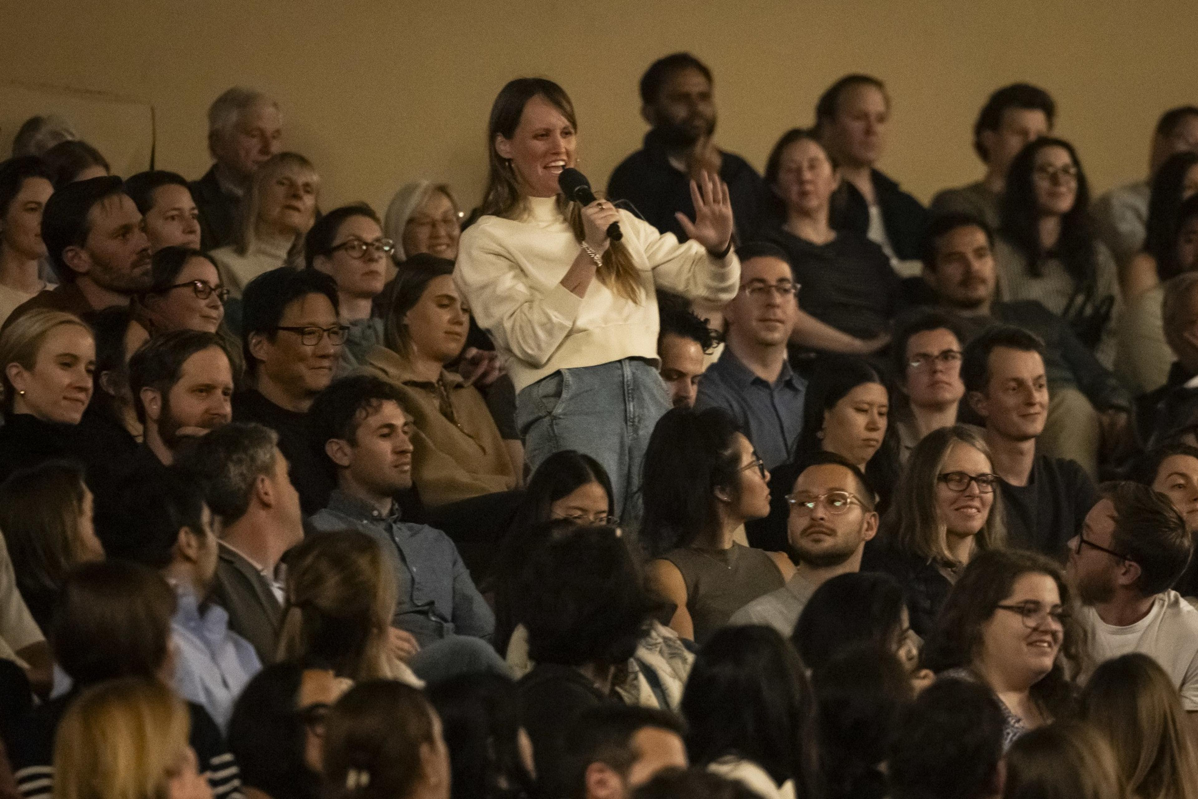 A woman in a light-colored sweater stands among a seated audience, holding a microphone and smiling as she addresses the crowd.