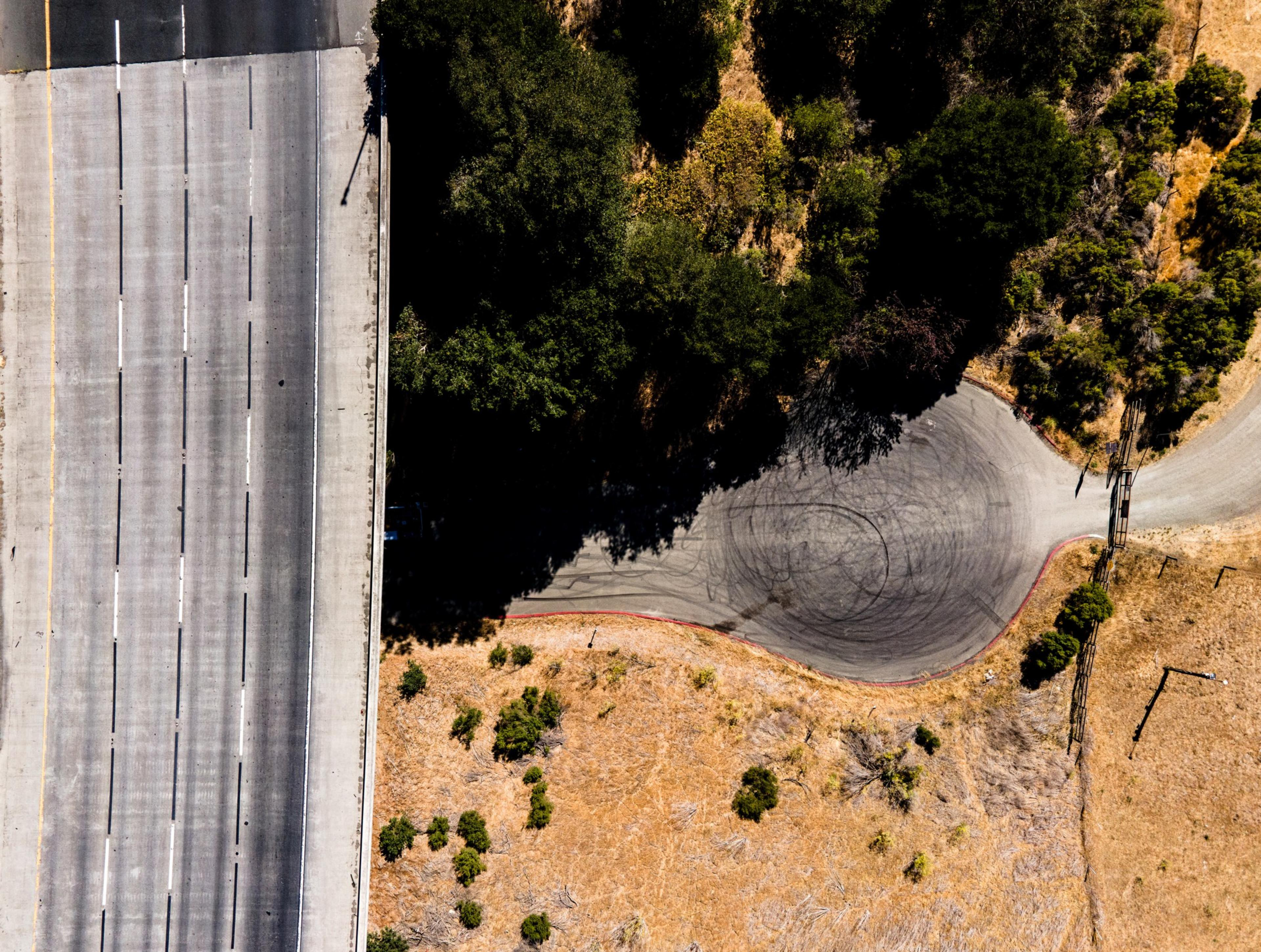 The image shows an aerial view of a highway on the left and a curved road on the right, surrounded by trees and dry grass, with visible skid marks.