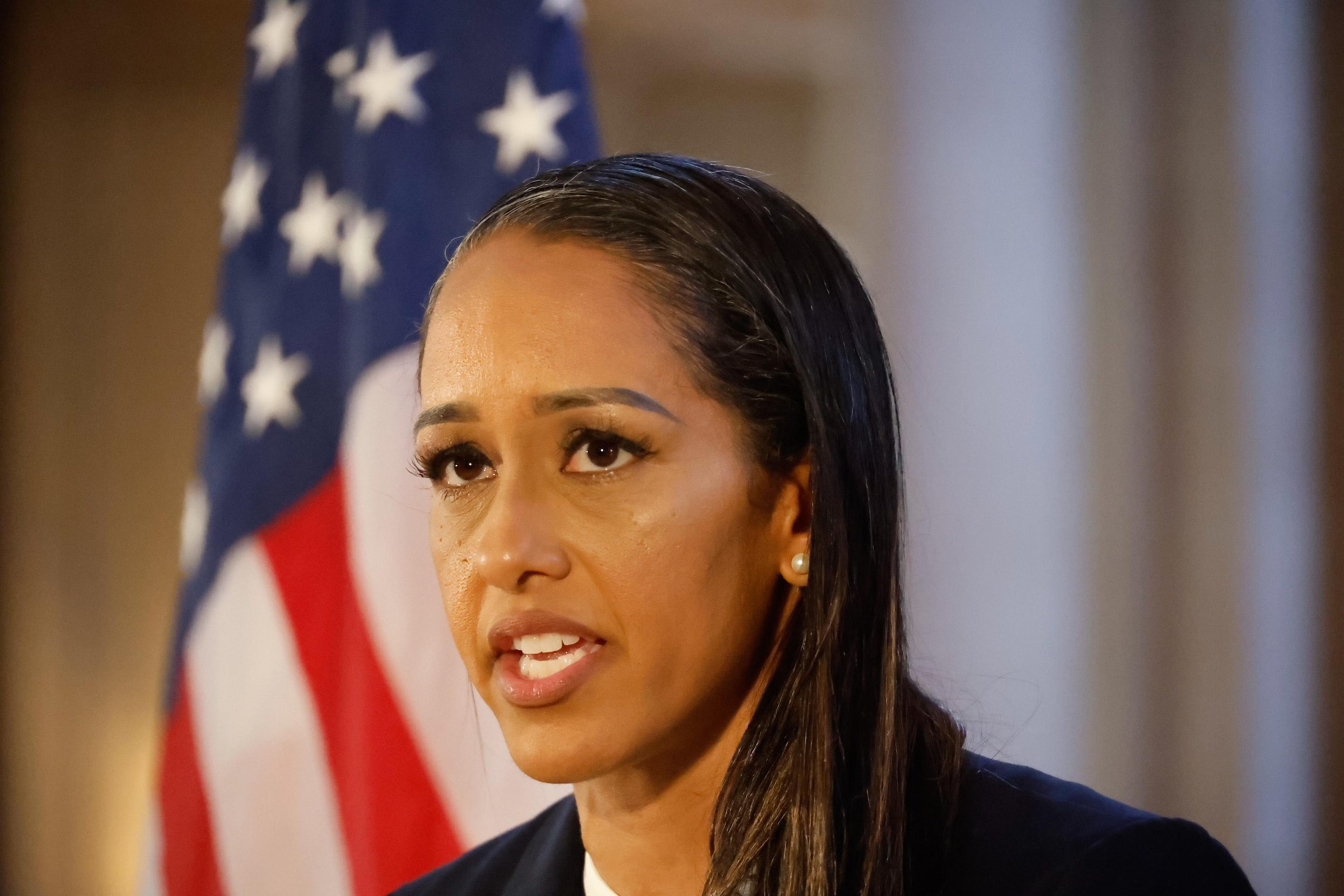 A woman with long dark hair speaks seriously, with an American flag blurred in the background.