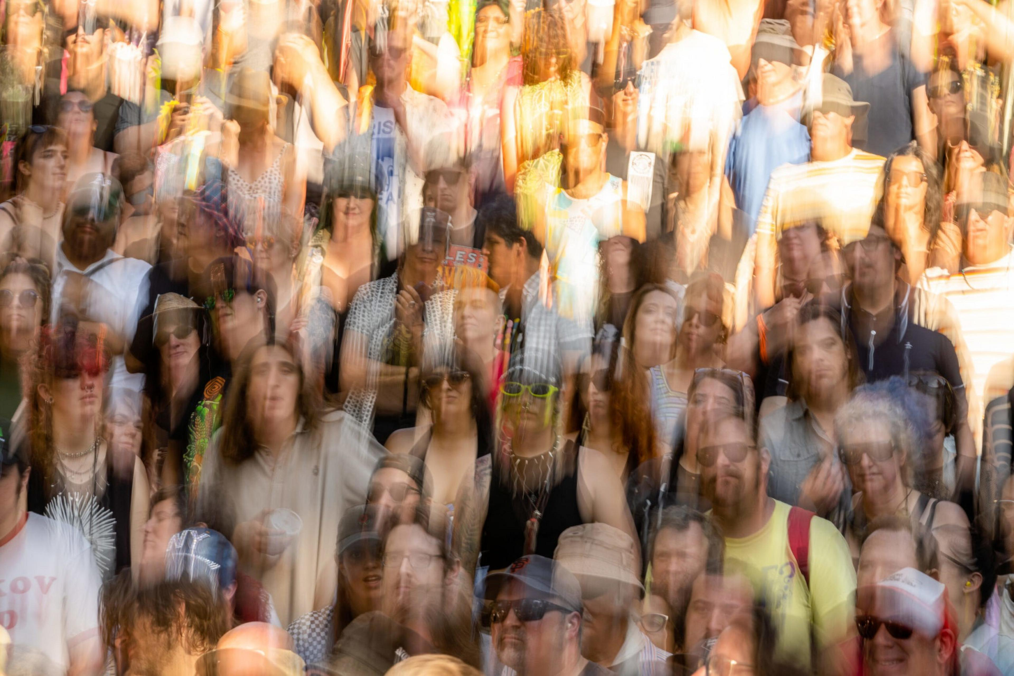 A crowd of people standing close together, many faces blurred by motion, with some wearing sunglasses and various clothing styles visible.