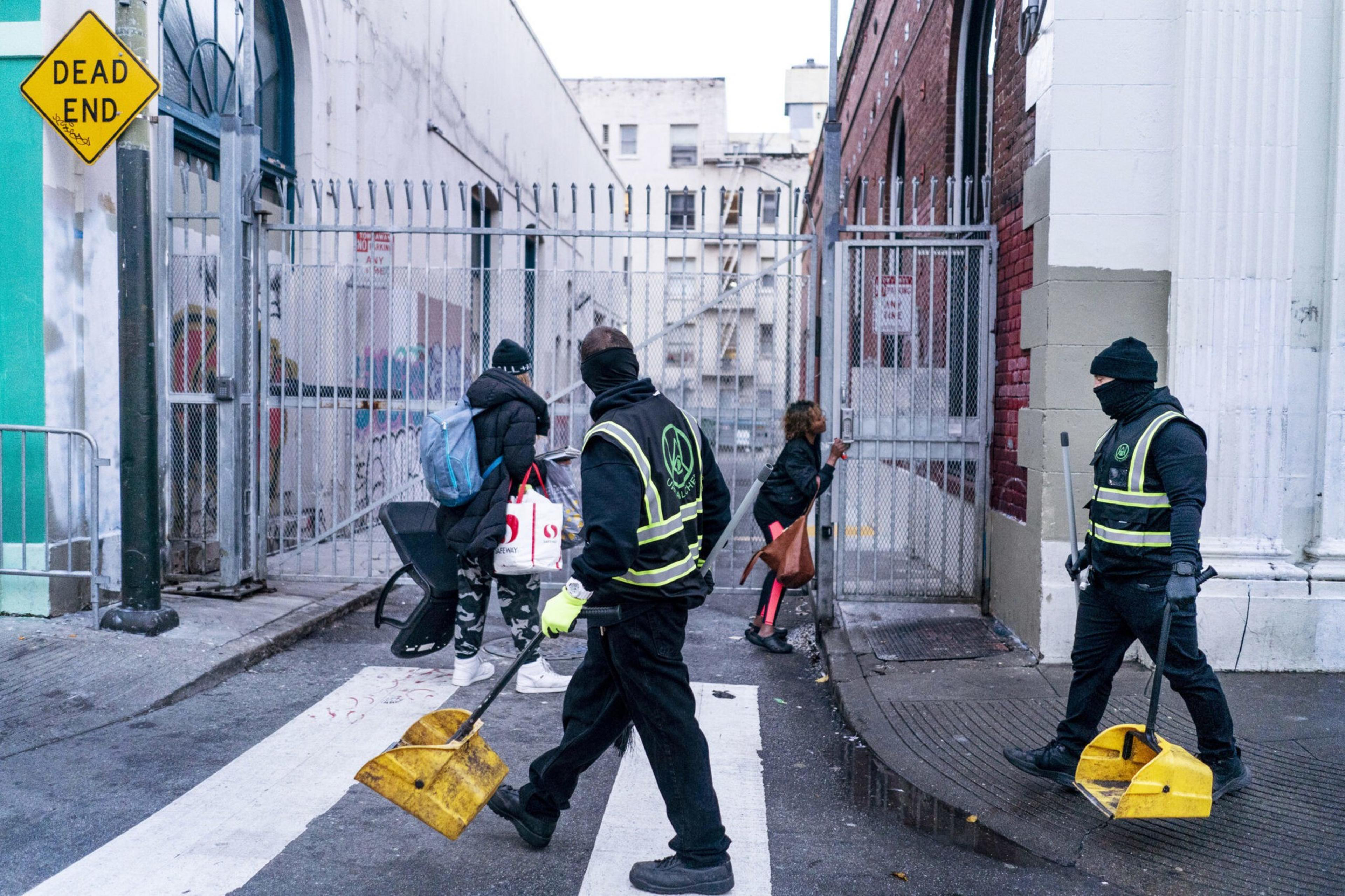 Three workers in safety vests, carrying shovels and gear, are in a narrow alley. A &quot;DEAD END&quot; sign is visible. Two people carrying bags are entering through a gate.