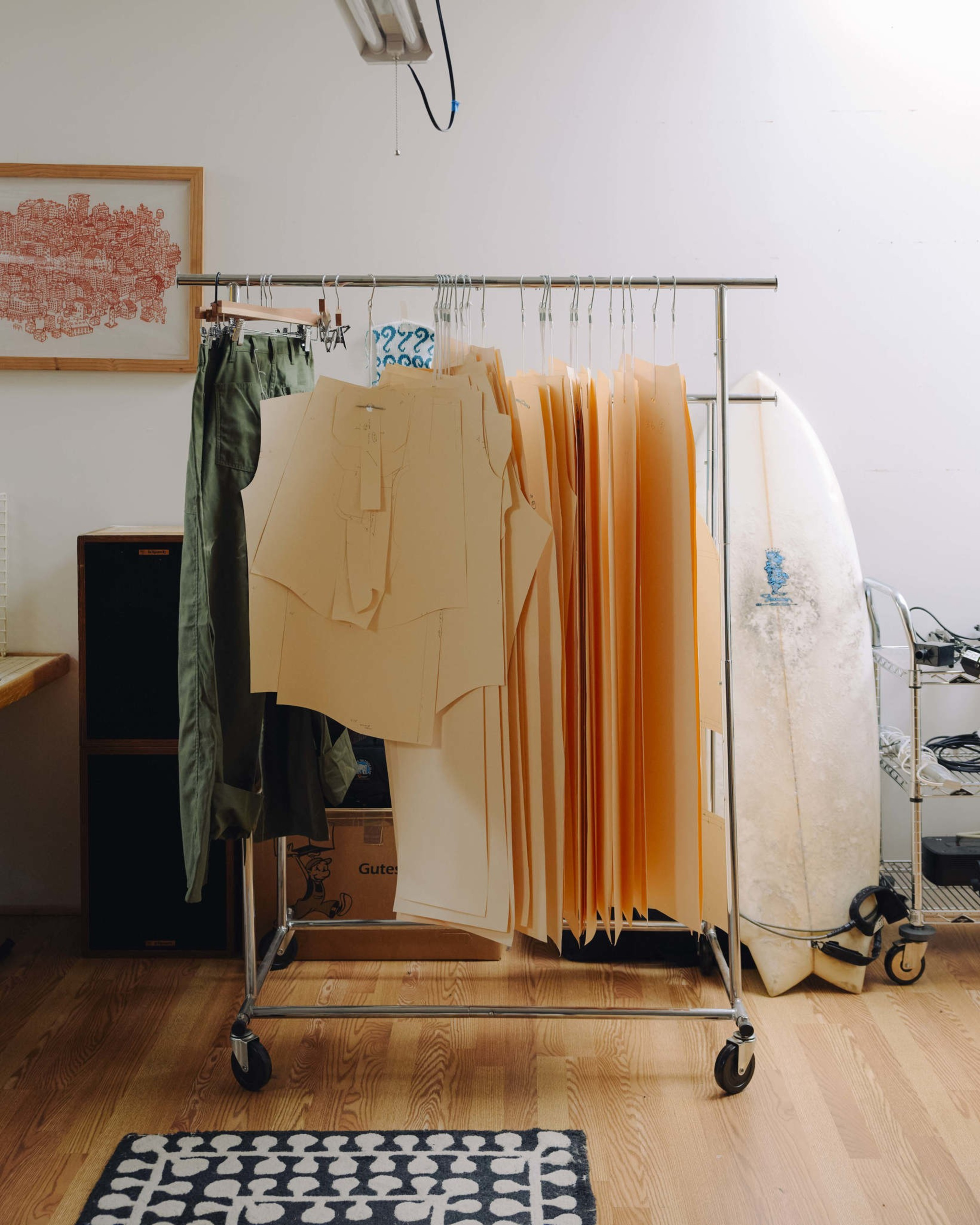 Clothing patterns, green pants, and a surfboard lean against the wall by a clothing rack on a wooden floor with a patterned rug nearby.
