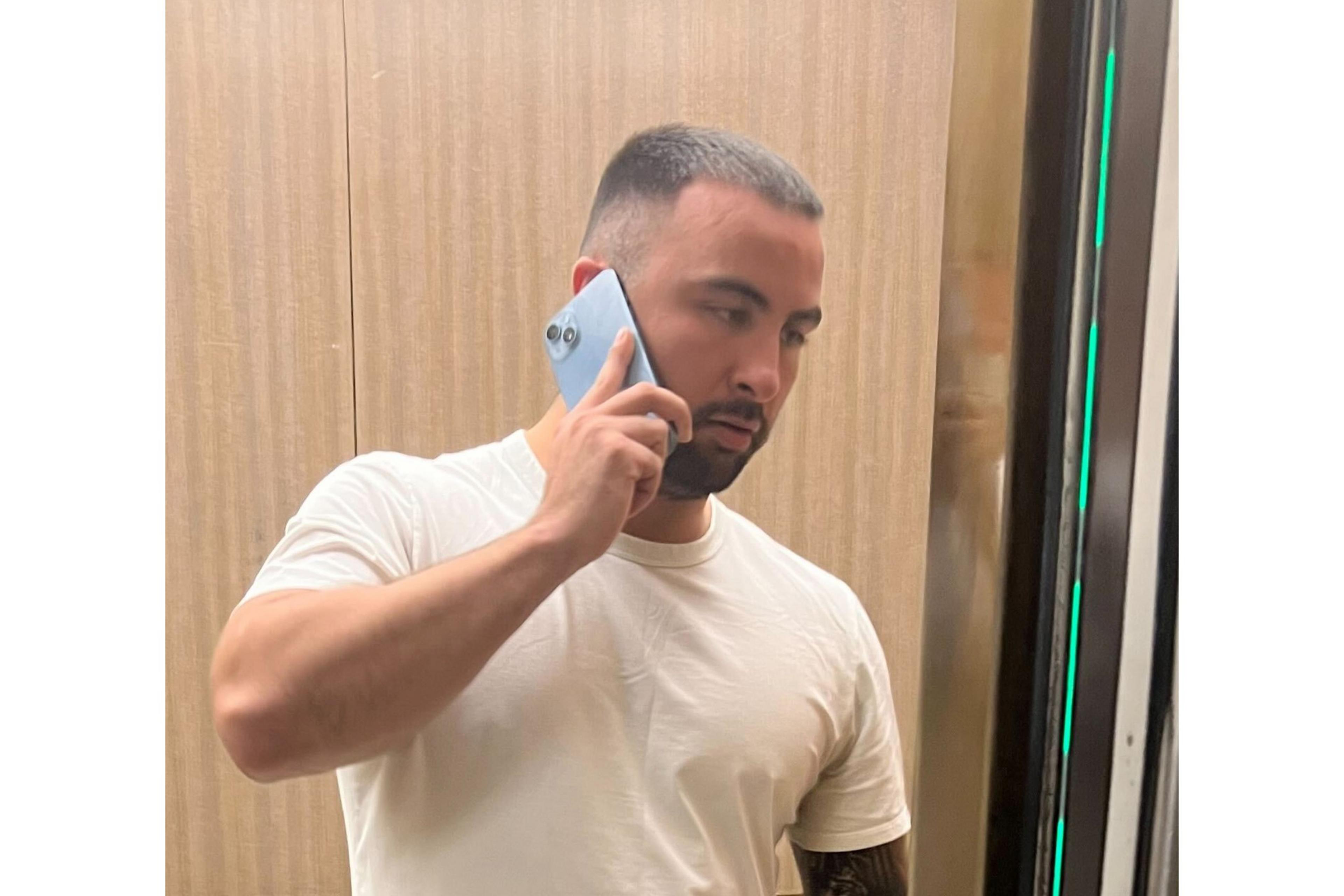 A man with short hair and a beard wears a white t-shirt while holding a blue smartphone to his ear, standing against a wood-paneled background.