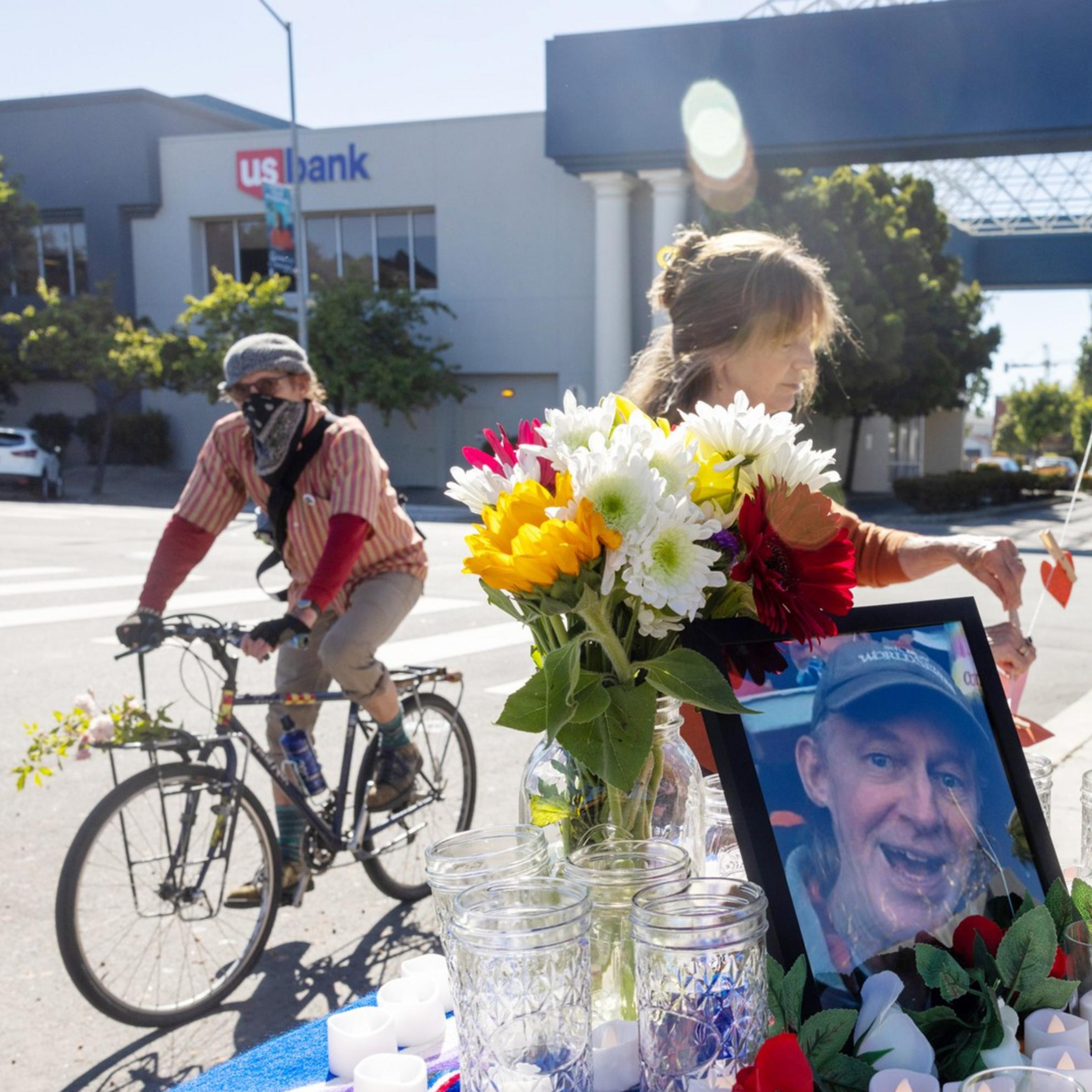 A memorial set on a city street with a photo, flowers, candles, and a colorful cloth atop a small table. A person bikes past.
