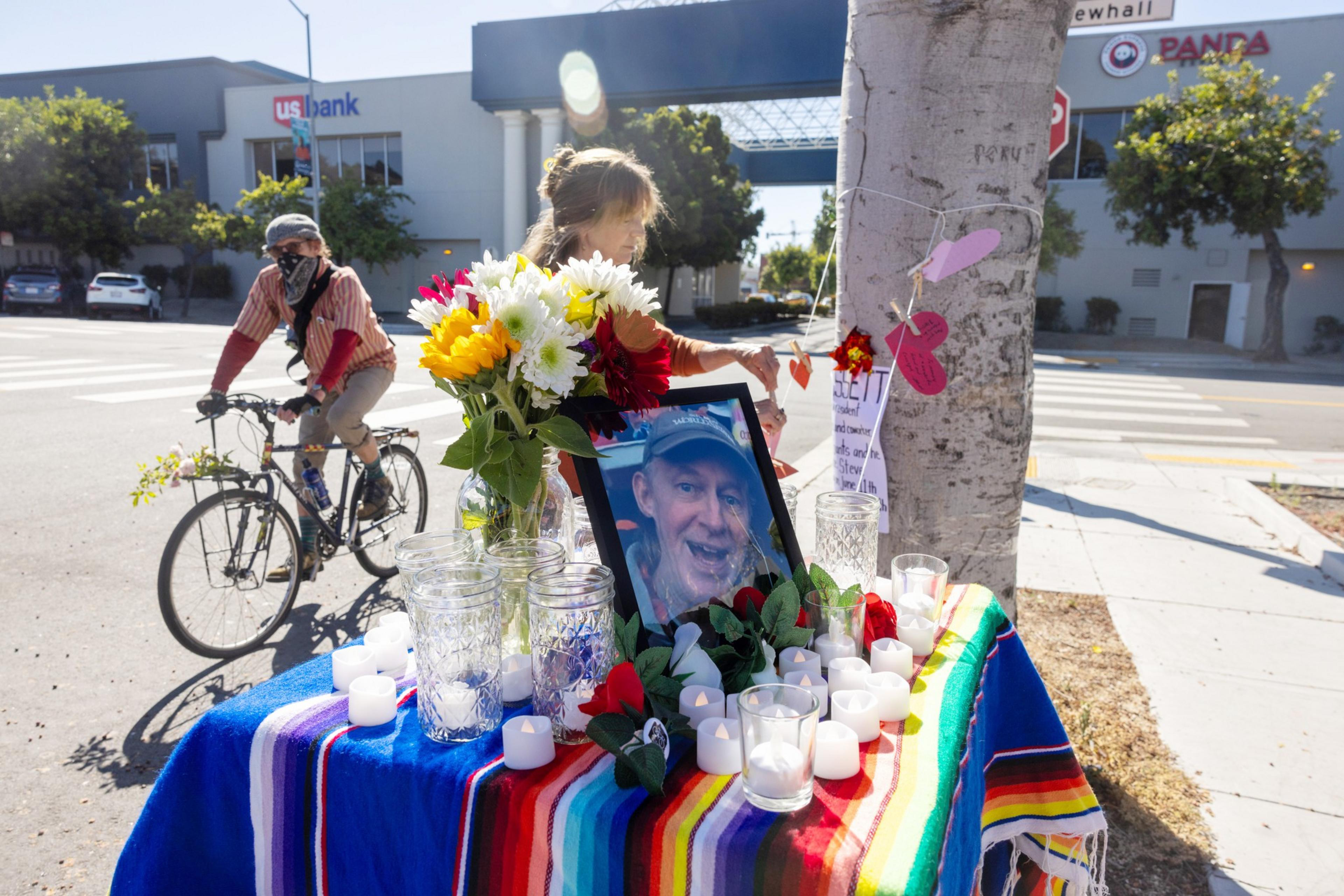 A memorial set on a city street with a photo, flowers, candles, and a colorful cloth atop a small table. A person bikes past.