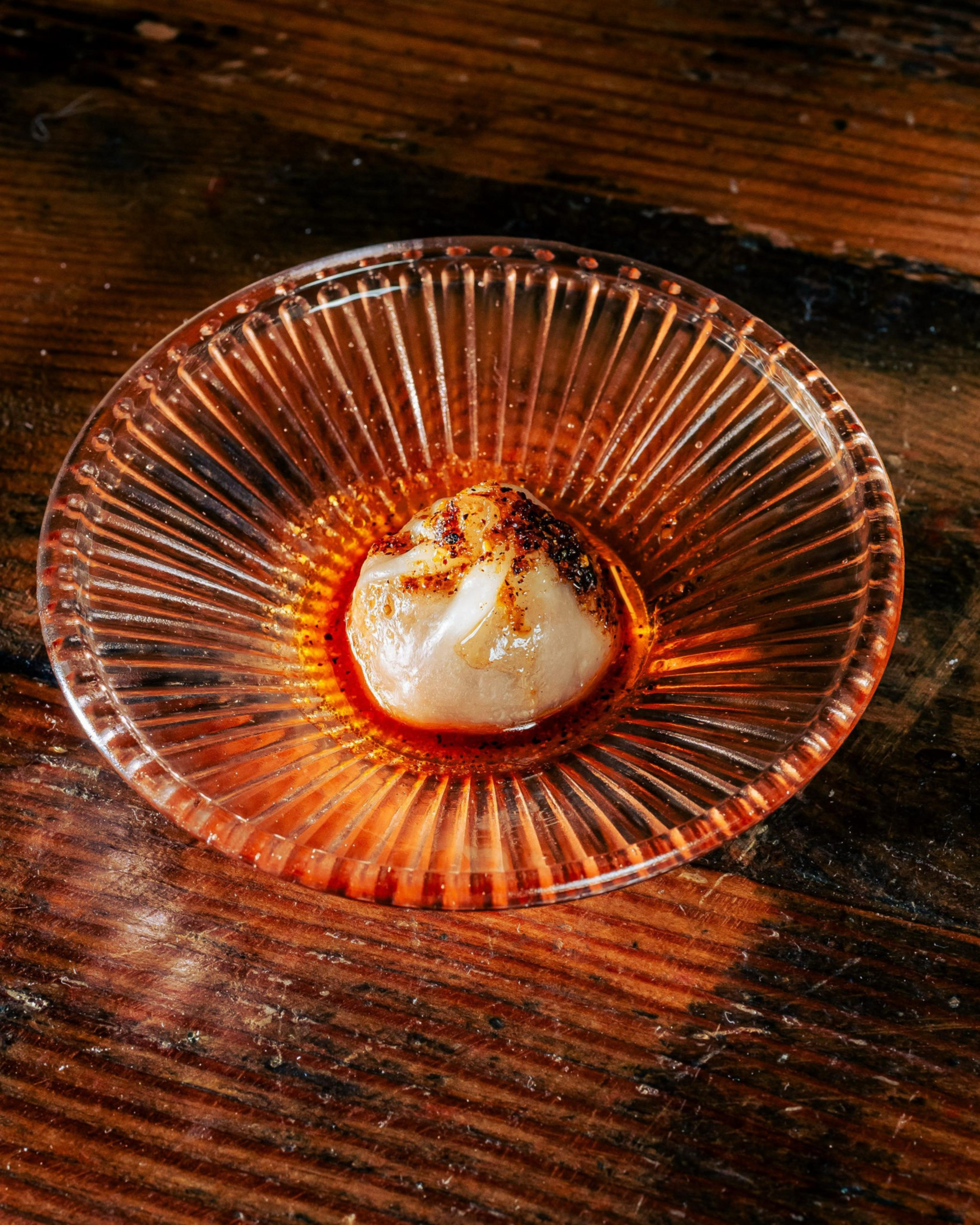 A single dumpling with a spicy seasoning sits on an orange, ridged glass plate, placed on a rustic wooden surface.