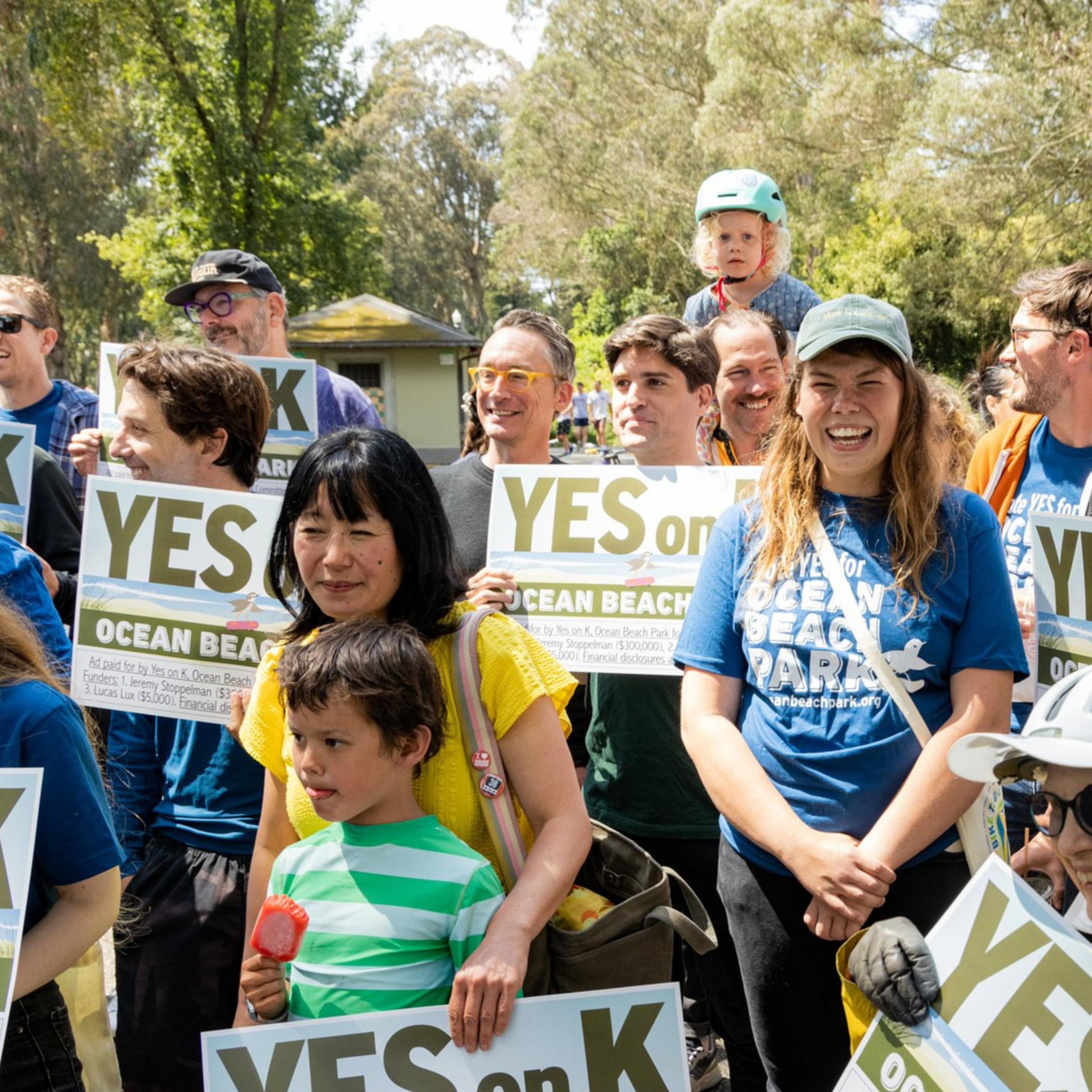 A diverse group of people, including children, stand outdoors holding signs that say &quot;YES on K&quot; and &quot;OCEAN BEACH PARK.&quot; They appear to be participating in a rally or gathering.