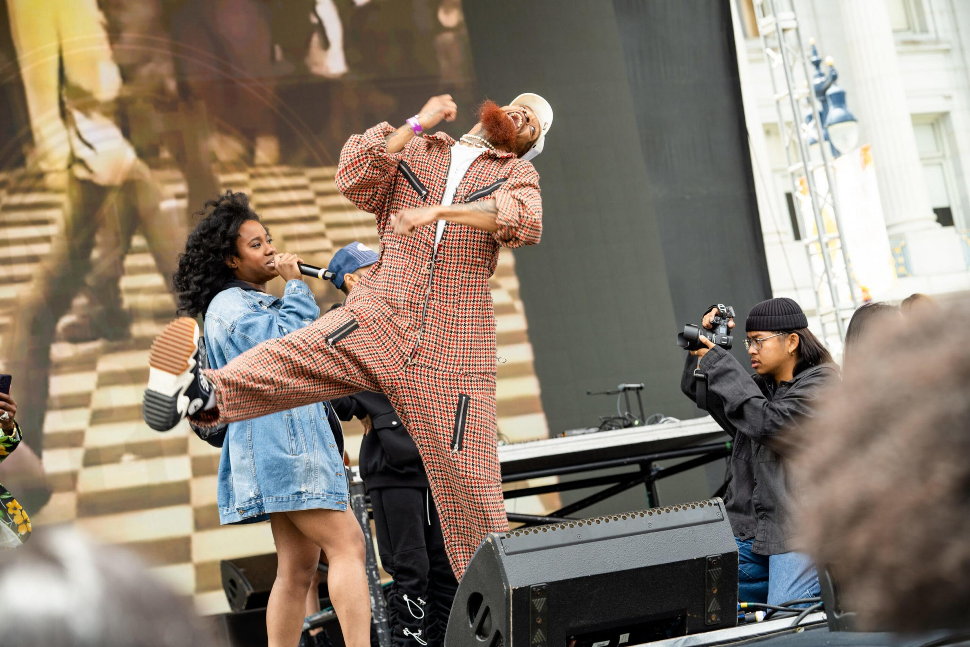 A man in a checkered jumpsuit jumps energetically on stage while a woman in a denim jacket holds a microphone and a photographer captures the moment.