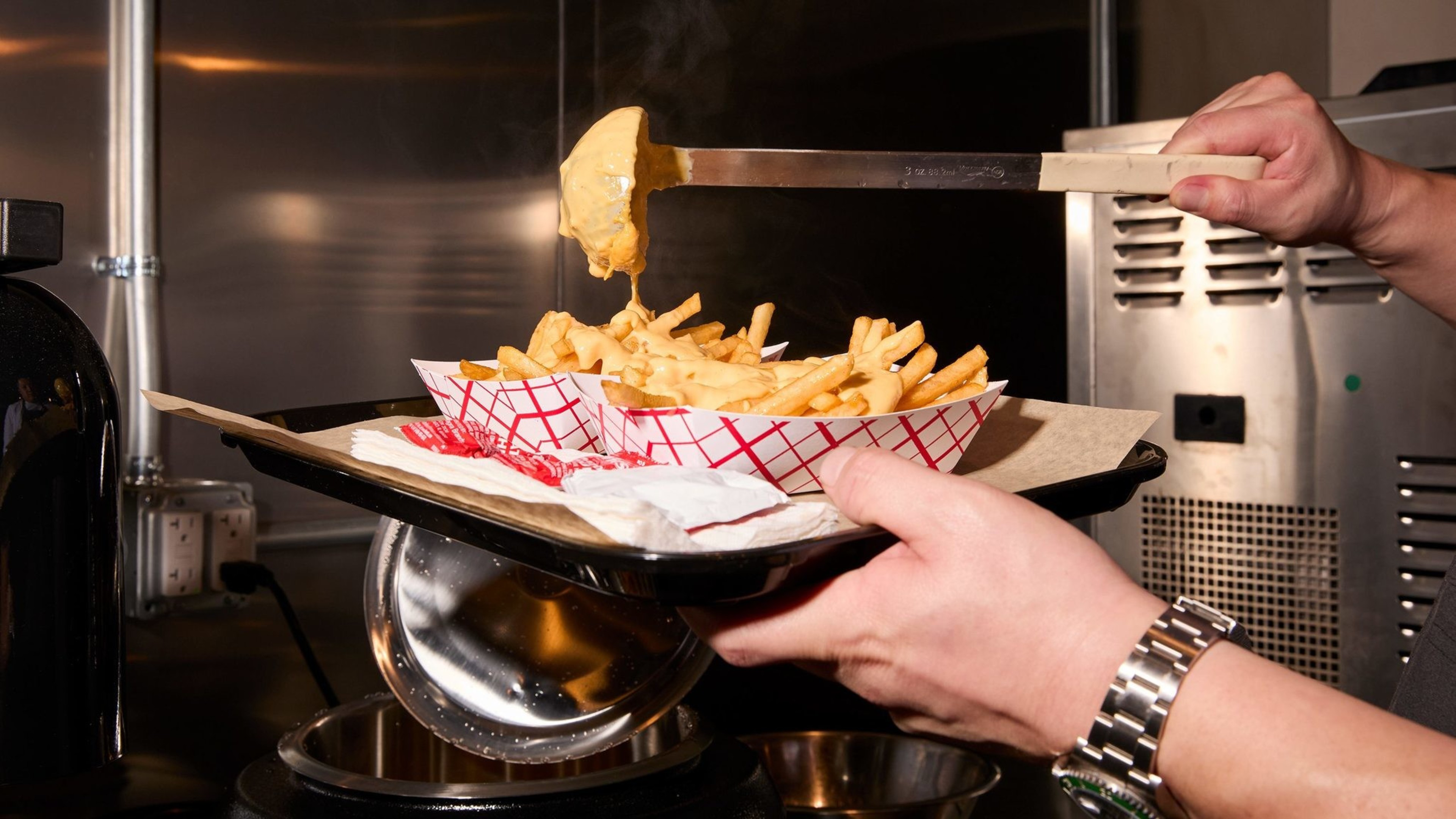 A hand pours melted cheese over a basket of french fries in a kitchen setting. The fries are in a red-and-white paper tray, and steam rises from the cheese.