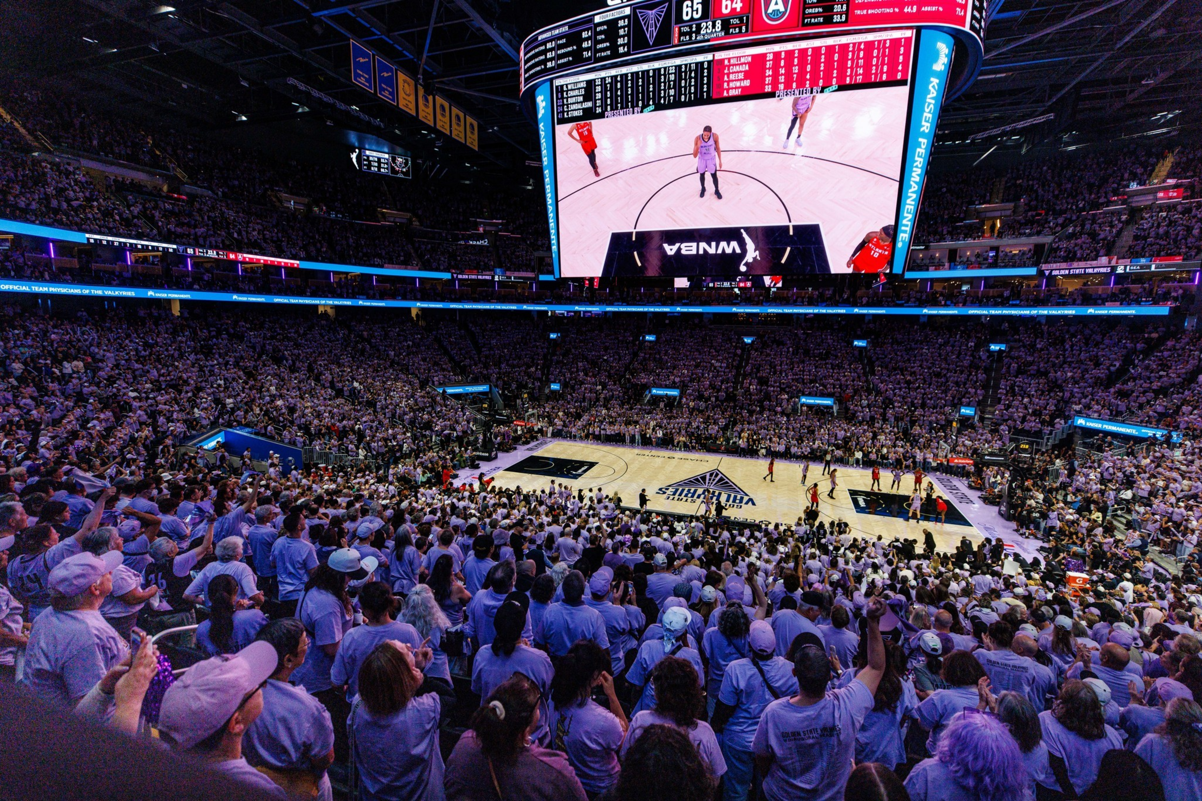 A packed indoor basketball arena with fans wearing purple watches a close WNBA game, the scoreboard shows 65-64 with under 24 seconds left.